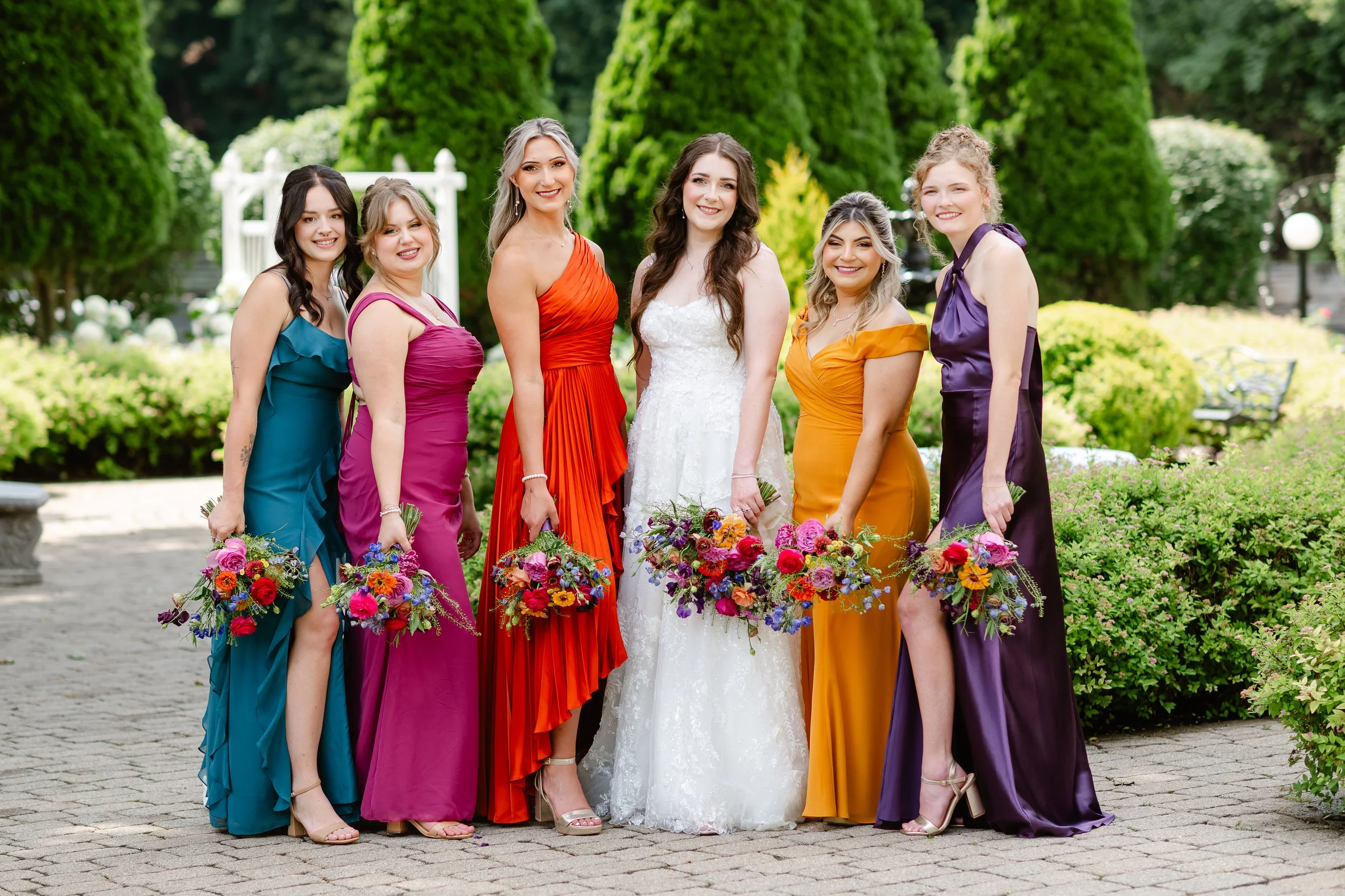 A group of seven women in colorful dresses, standing outdoors in a garden setting, each holding a bouquet of flowers, celebrating a special occasion.
