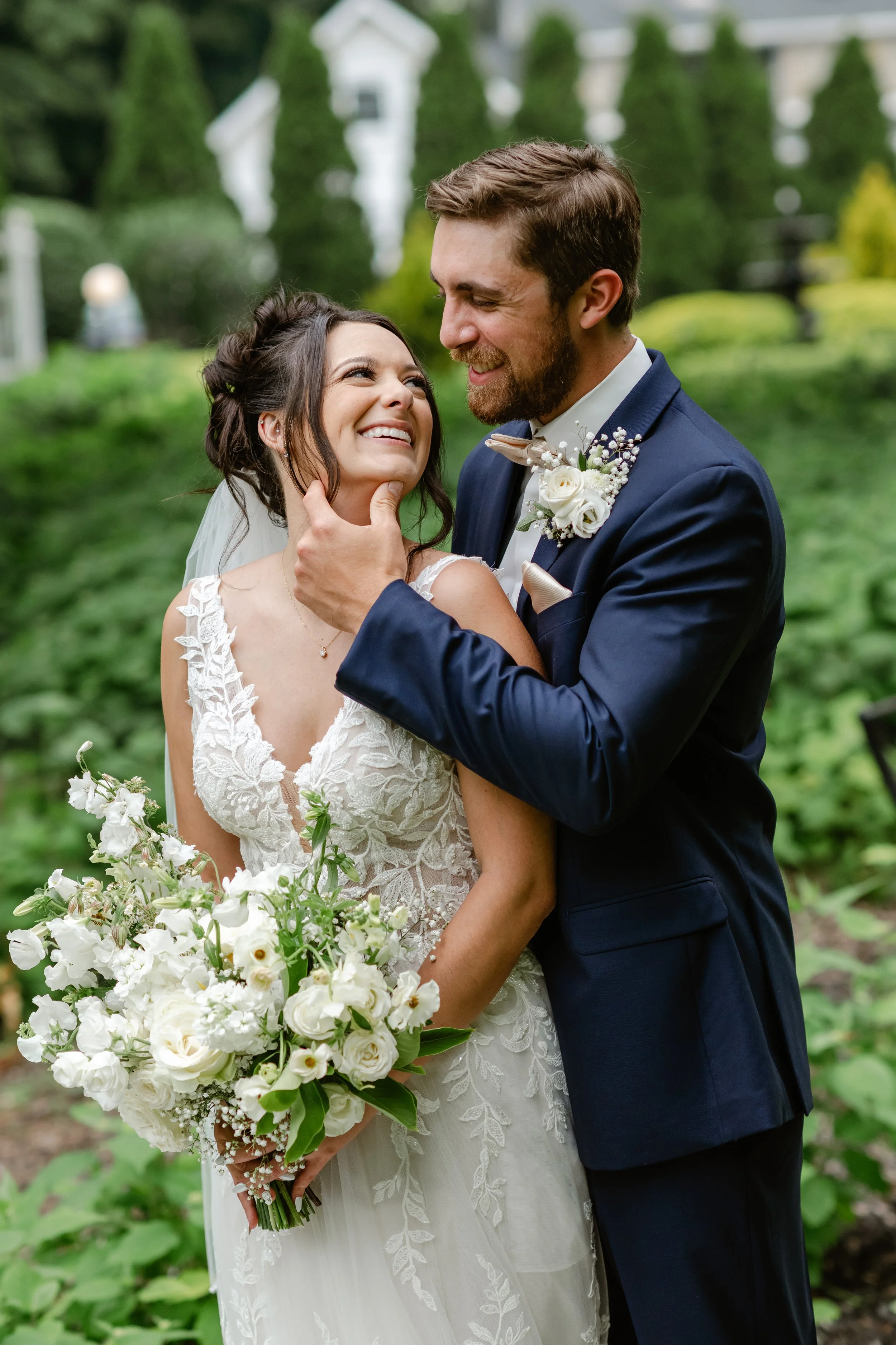 A bride and groom sharing a joyful moment outdoors, with the groom gently holding the bride's chin. The bride is holding a large bouquet of white flowers, and the couple is dressed in wedding attire with greenery in the background.