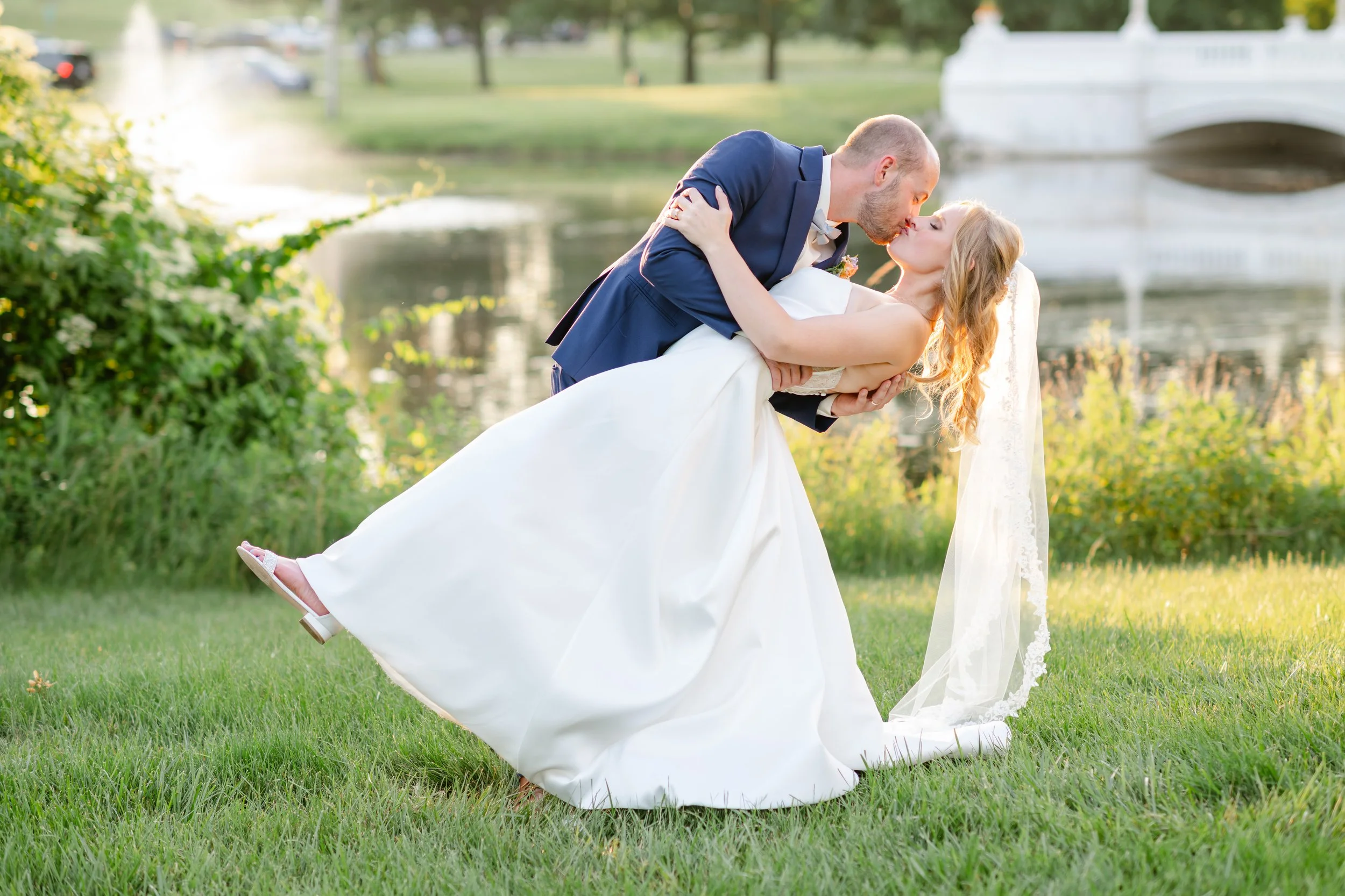 A newlywed couple, the groom in a navy suit and the bride in a white wedding gown with a veil, are outdoors by a lake or pond. The groom is dipping and kissing the bride, who is leaning back in his arms on a grassy area with trees and a stone bridge 