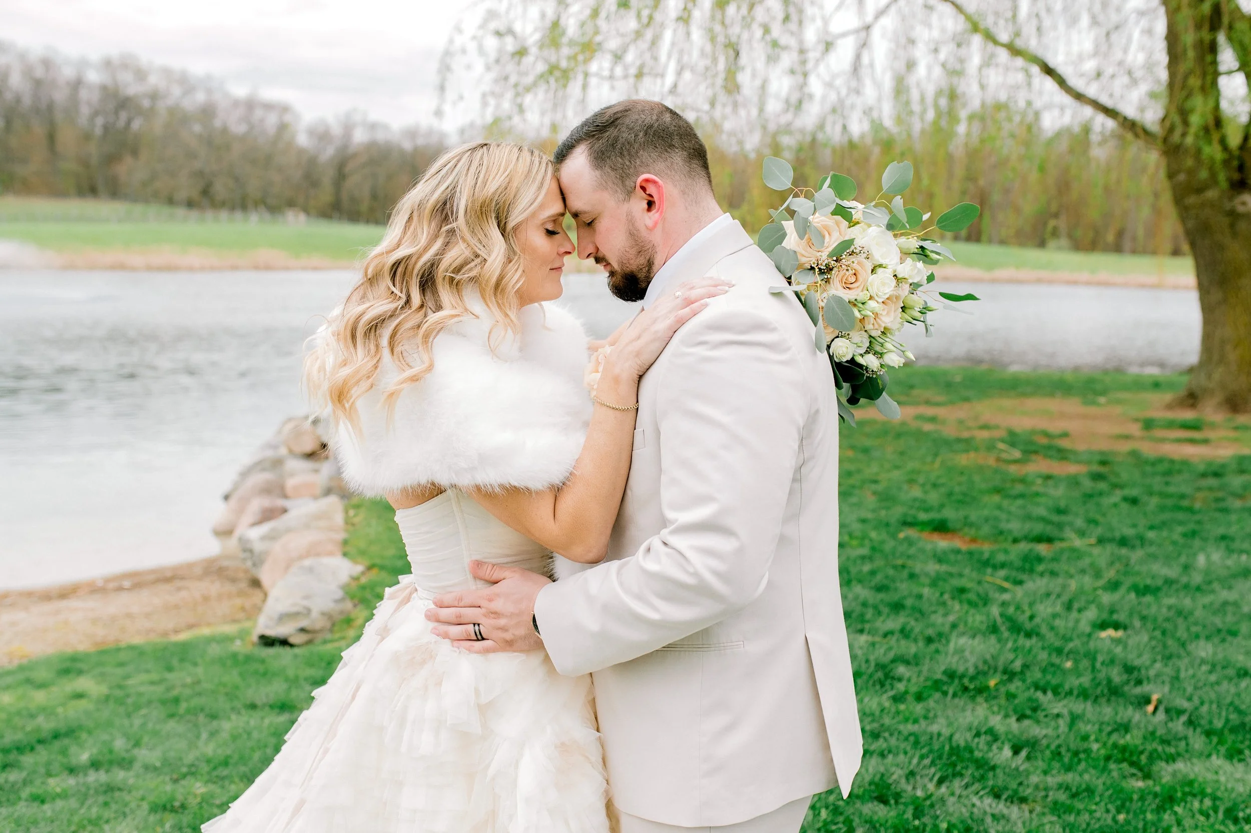 Bride and groom stand close together outdoors near a river, with their foreheads touching and eyes closed. The bride has long, wavy blonde hair and wears a cream-colored wedding dress with a white fur stole. The groom has short brown hair and a beard
