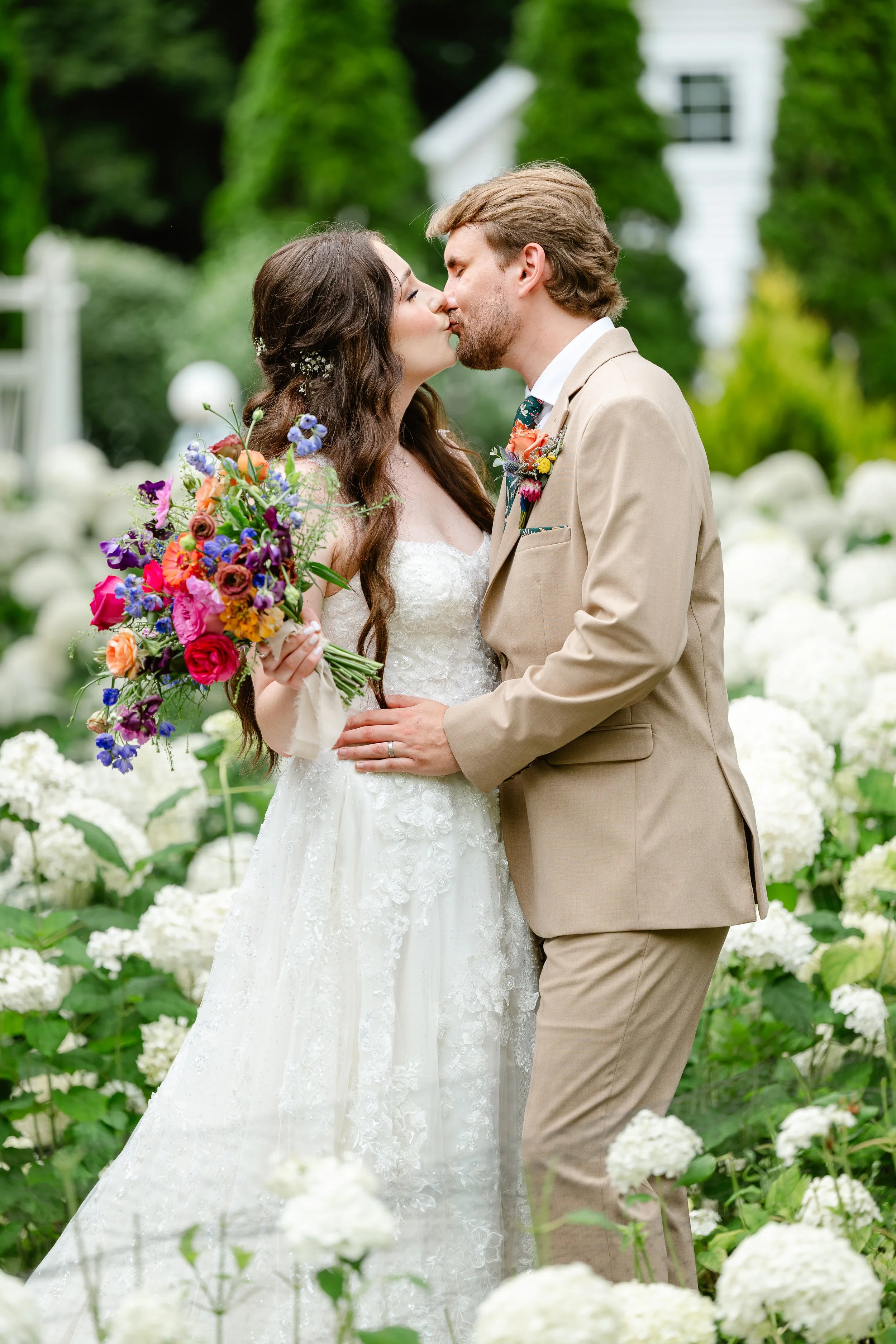 A bride and groom kiss outdoors among white hydrangeas, with the bride holding a colorful bouquet, wearing a white lace wedding gown and the groom in a tan suit.