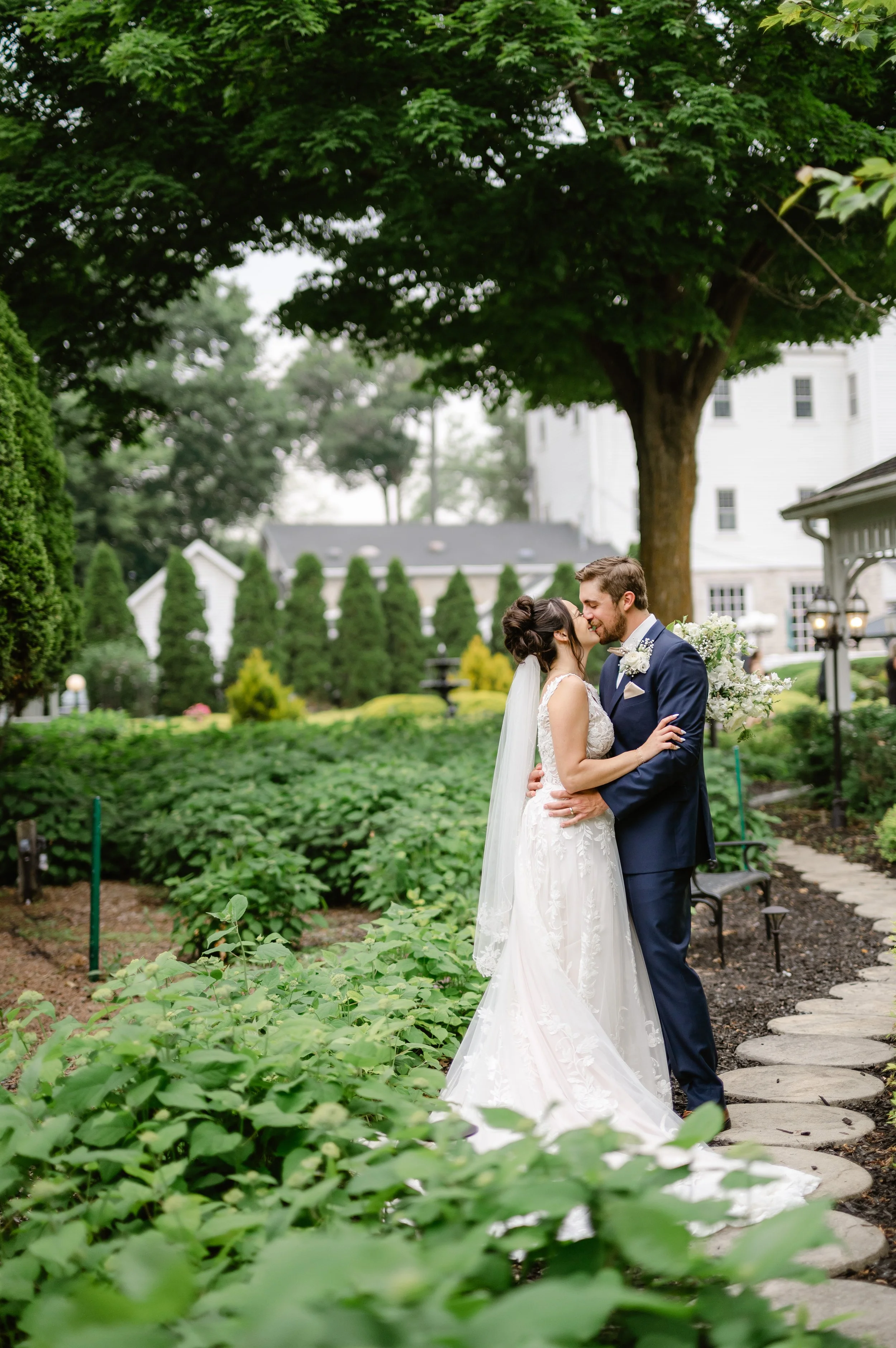 A newlywed couple sharing a kiss in a lush garden with green bushes and tall trees in the background.
