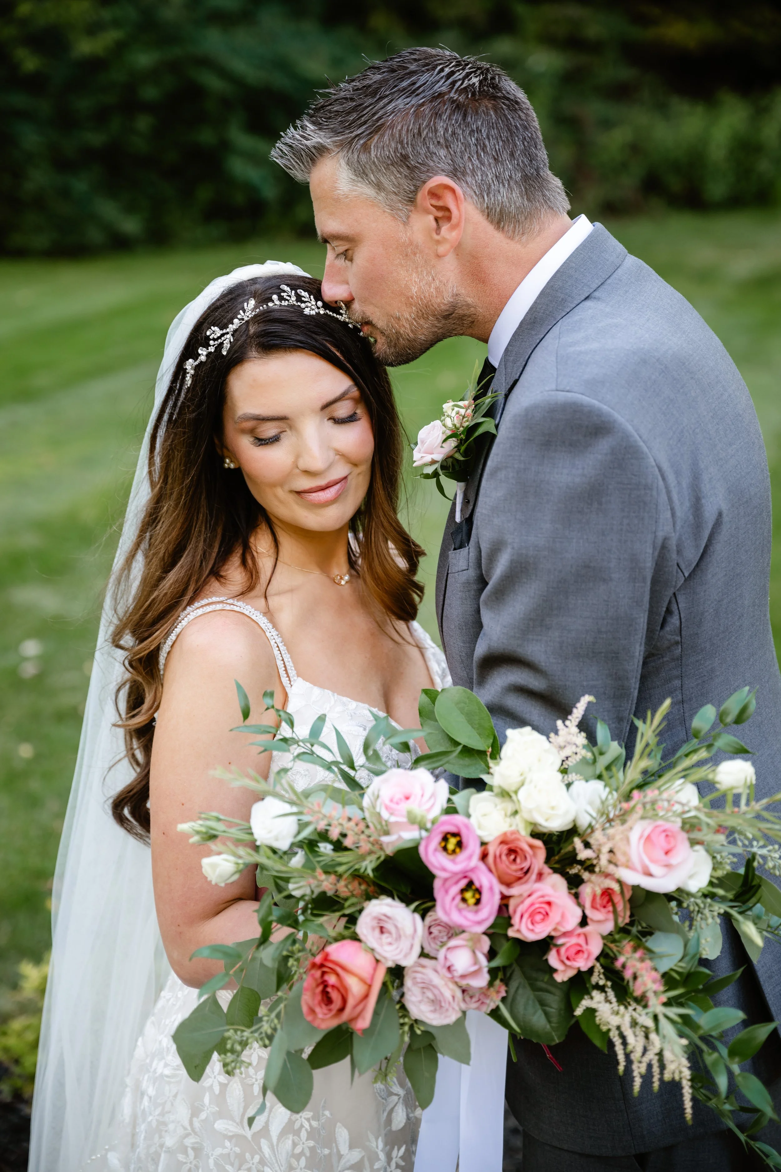 A newlywed couple outdoors; the groom kisses the bride on the forehead. The bride wears a white wedding dress with lace straps, a veil, and a floral headpiece. She holds a large bouquet of pink, white, and peach roses with greenery. The groom wears a