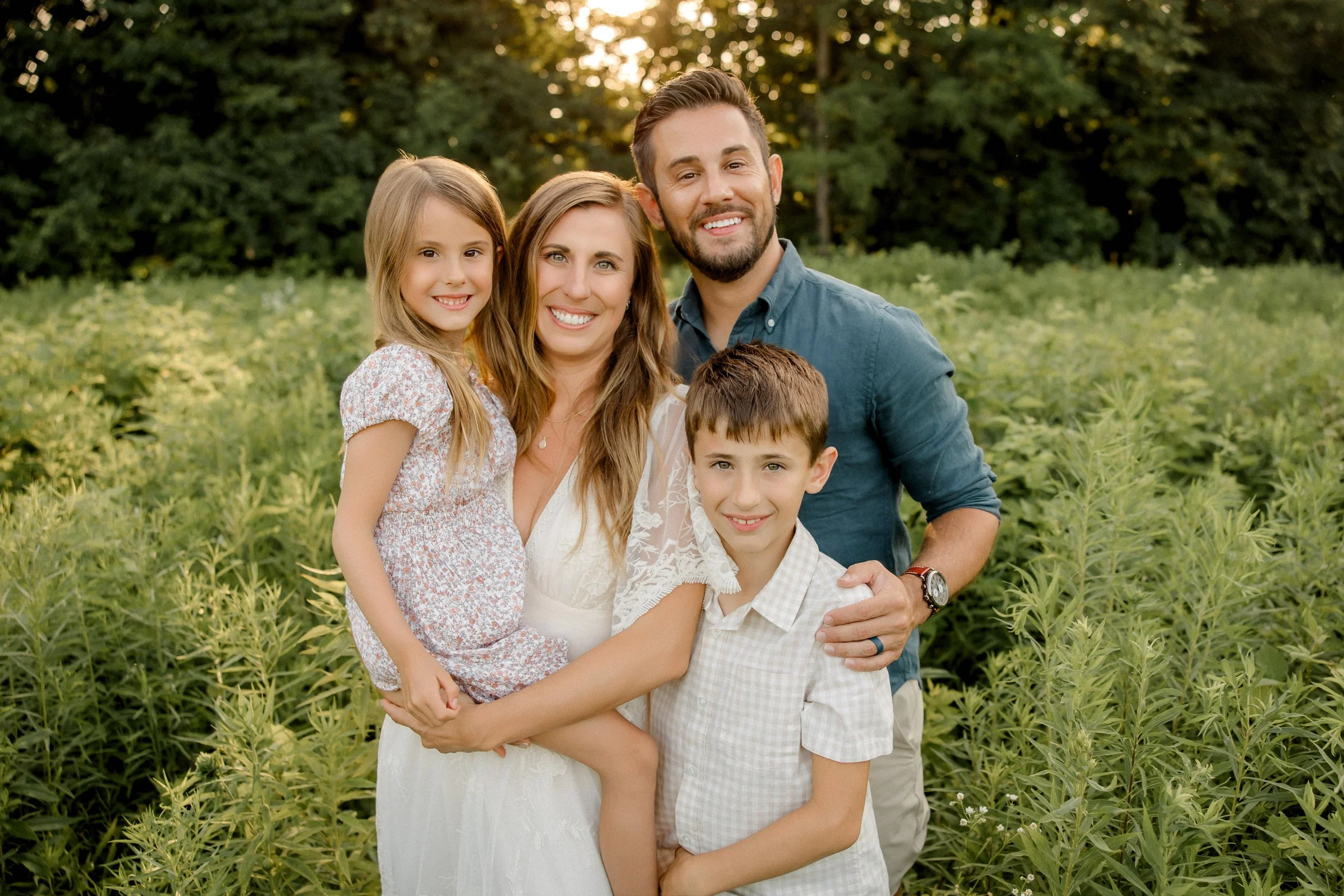 A family of four standing in a green field with trees in the background, smiling at the camera during sunset.