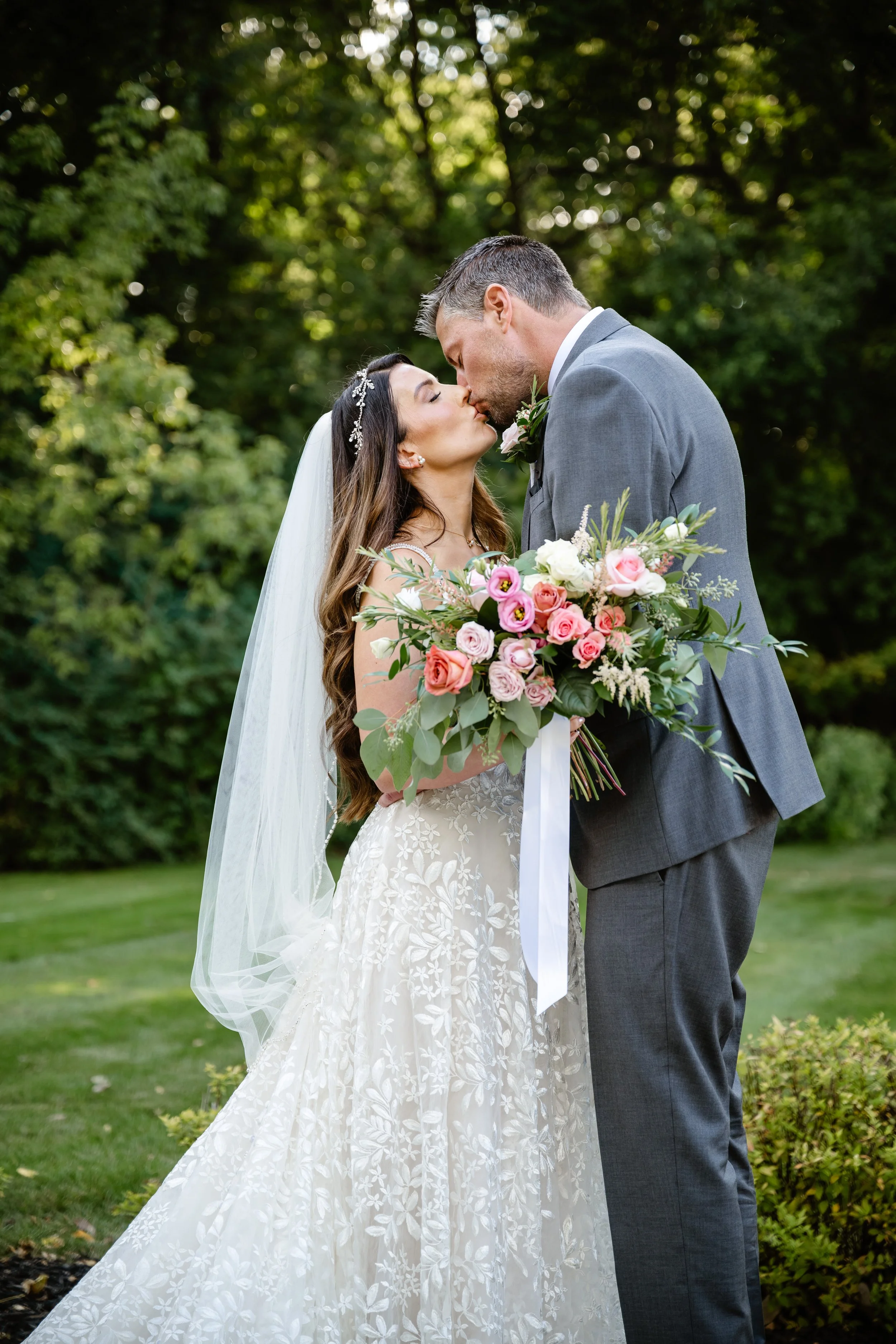 A bride and groom kissing outdoors during wedding, with the bride holding a bouquet of pink and white flowers, and a green, wooded background.