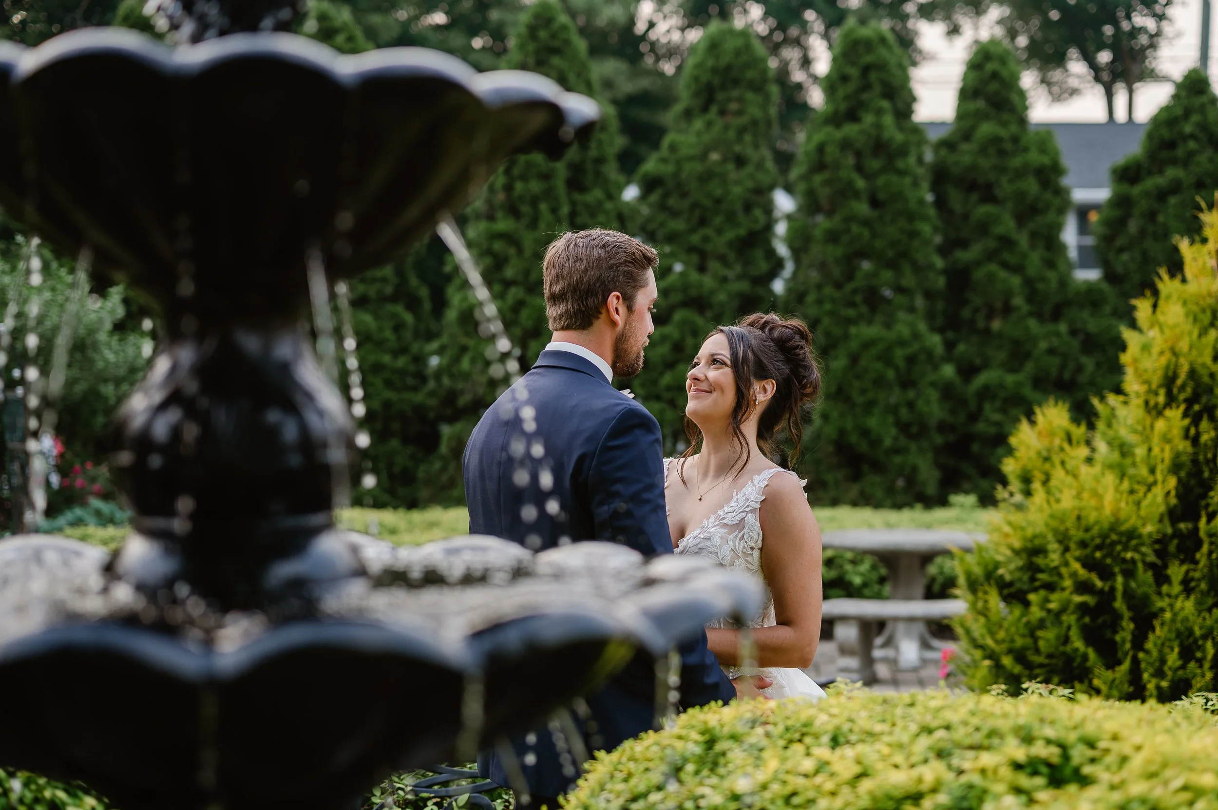 A bride and groom standing close together in a garden, smiling and gazing into each other's eyes, with greenery and trees in the background.