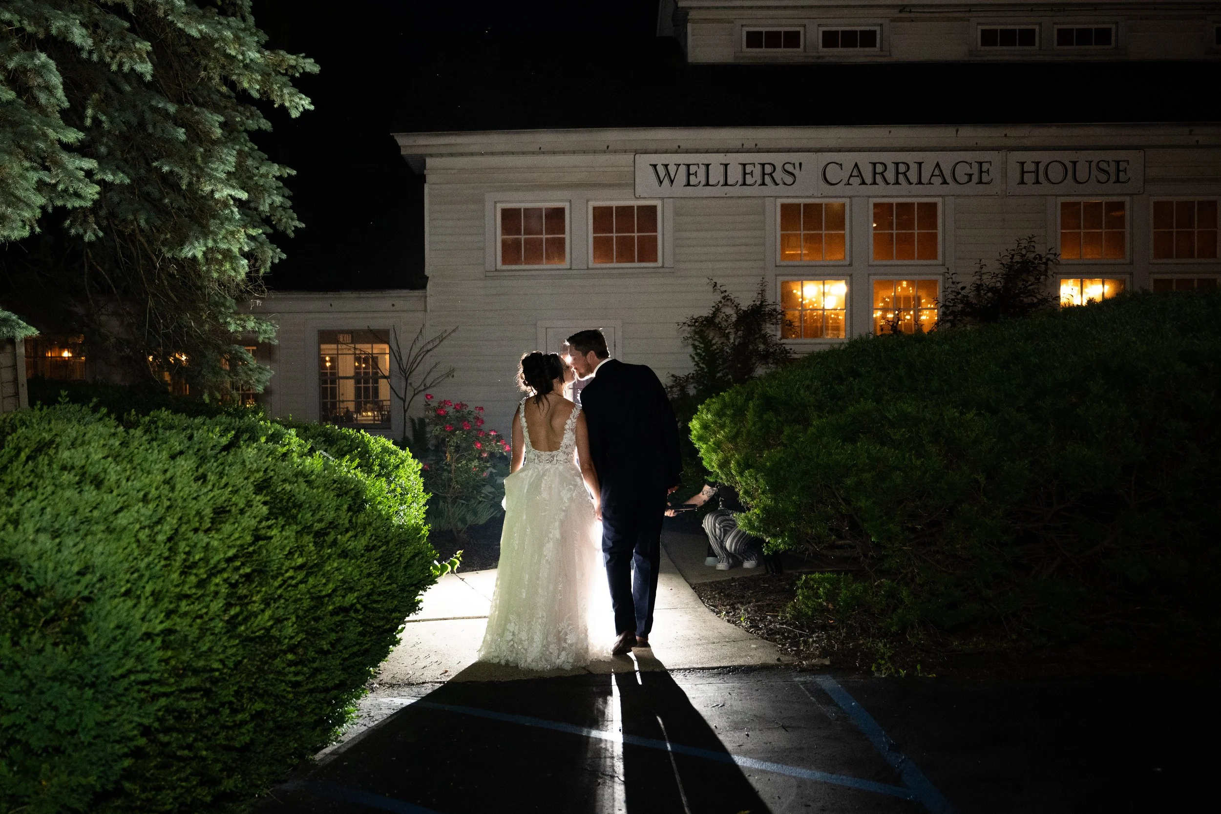 A bride and groom walking hand in hand at night outside Wellers' Carriage House, illuminated by surrounding lights, with lush bushes and a large tree nearby.