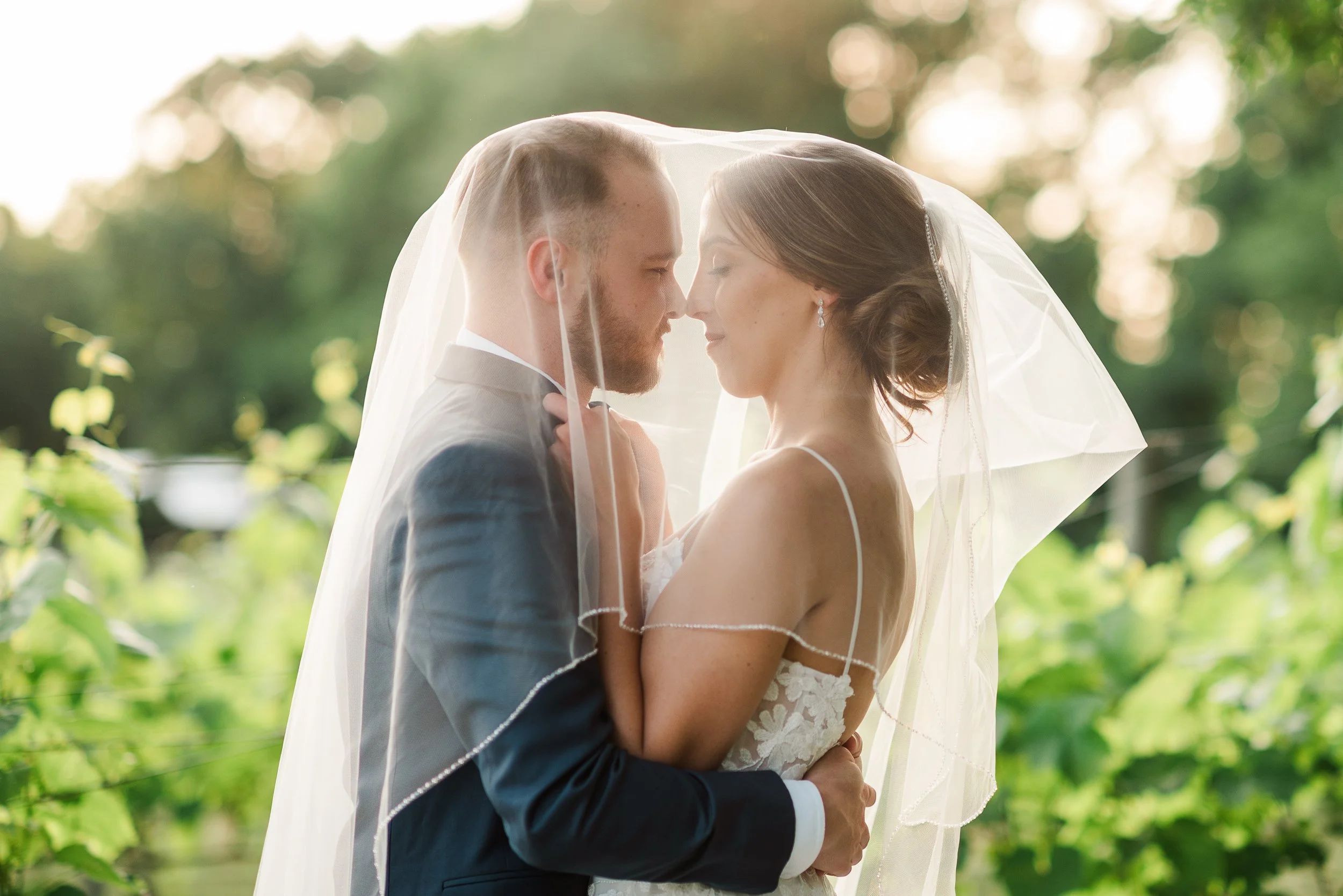 A bride and groom standing close together, under a bridal veil, outdoors with greenery in the background, sharing an intimate moment.