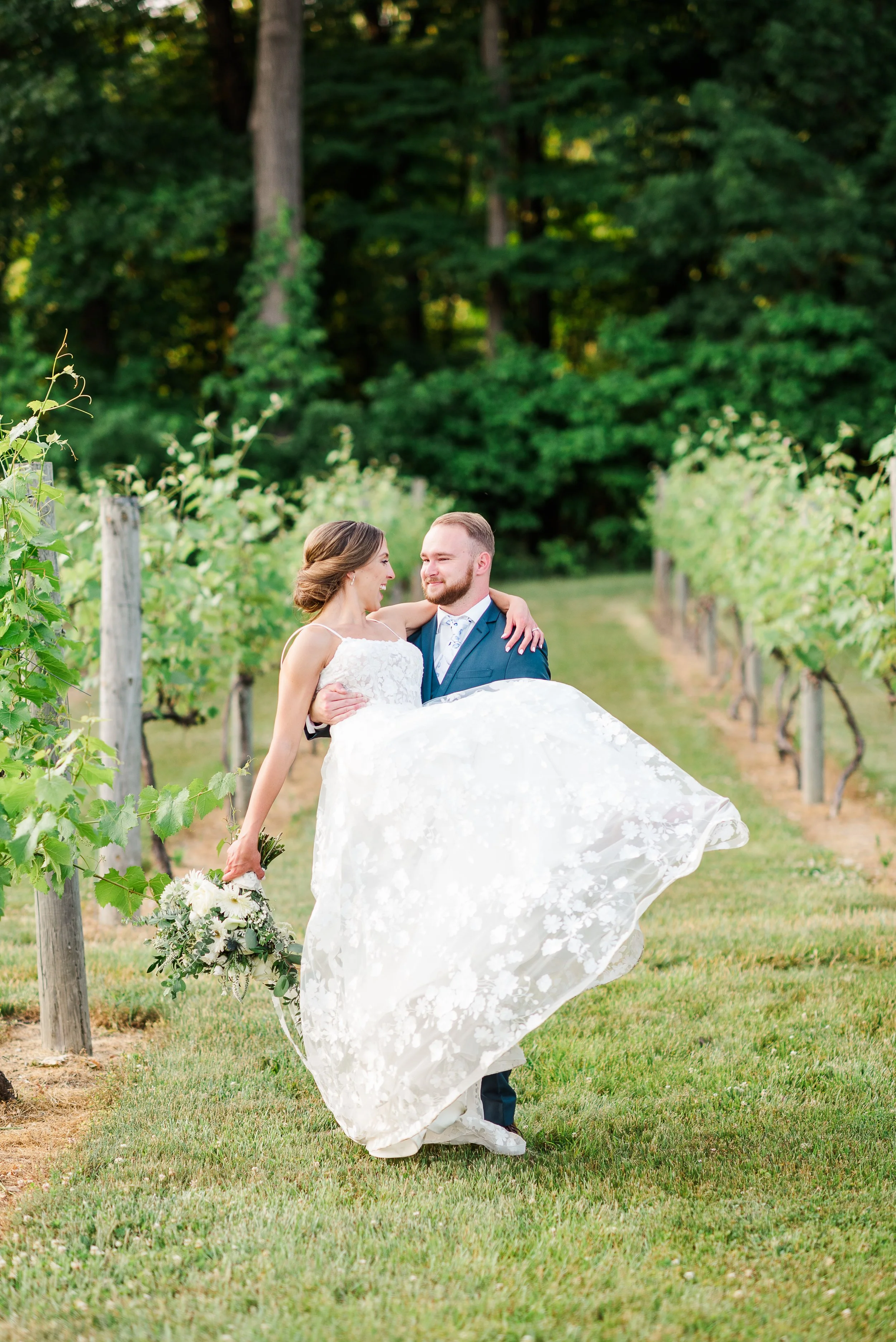 A newlywed couple in wedding attire smiling and embracing in a vineyard, with the groom carrying the bride, who is holding a bouquet.