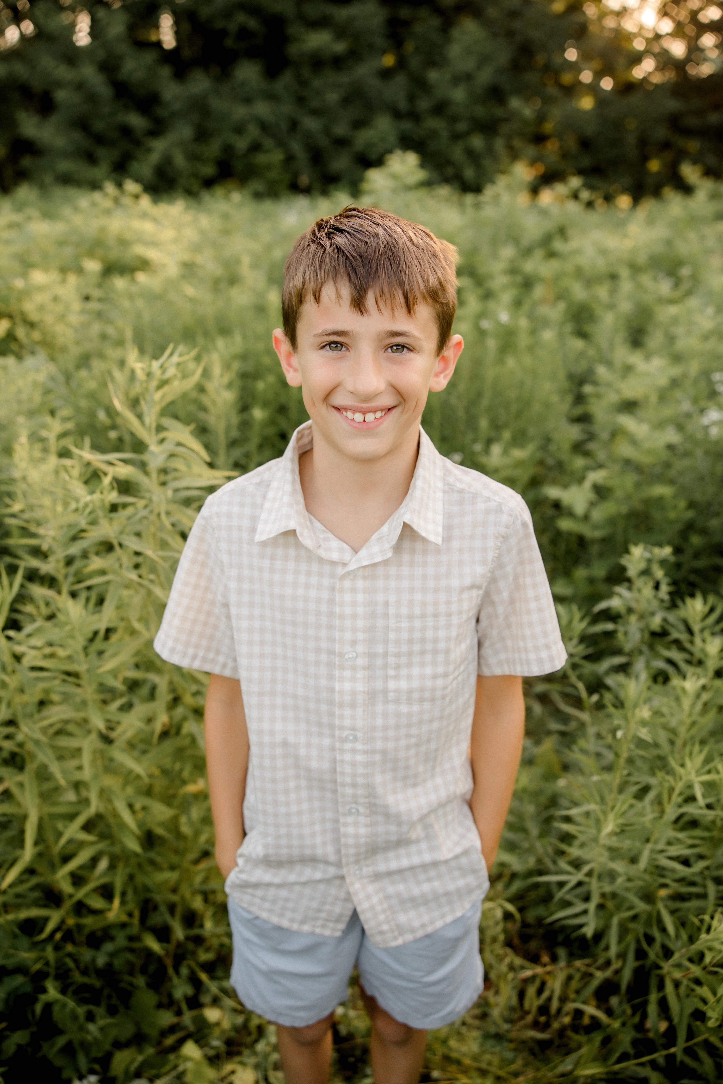 A young boy with brown hair and blue eyes smiling outdoors in a green, leafy setting, wearing a light-colored checkered short-sleeve shirt and gray shorts.