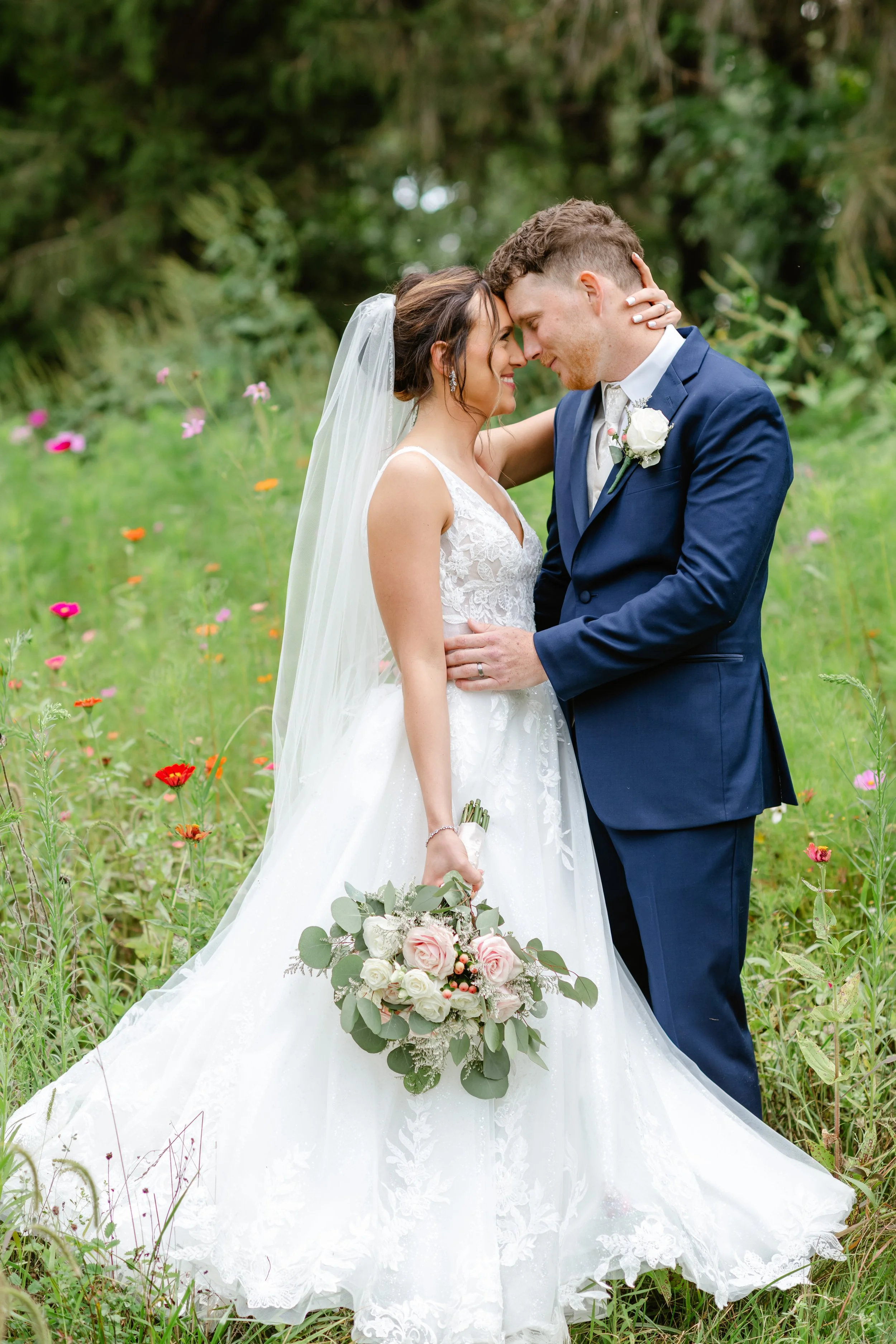 A bride and groom in a field of wildflowers, embracing and holding hands. The bride wears a white lace wedding dress with a veil and holds a bouquet of pink and white roses. The groom wears a navy blue suit with a white shirt and boutonniere.