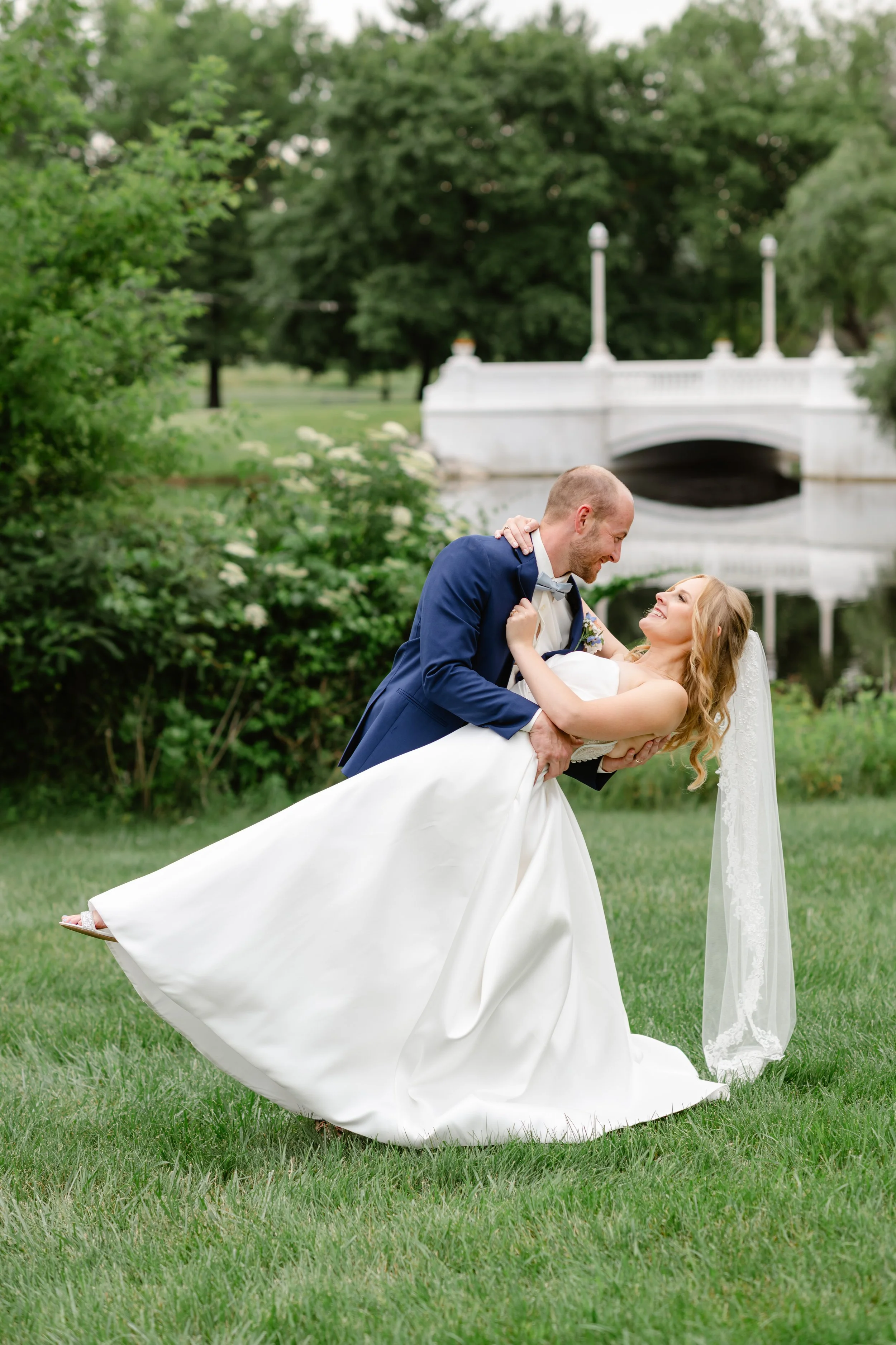 A newlywed couple dancing outdoors on a grassy area with a small bridge and lush green trees in the background. The groom is wearing a navy blue tuxedo and the bride is in a white wedding gown with a veil, smiling and looking at each other.