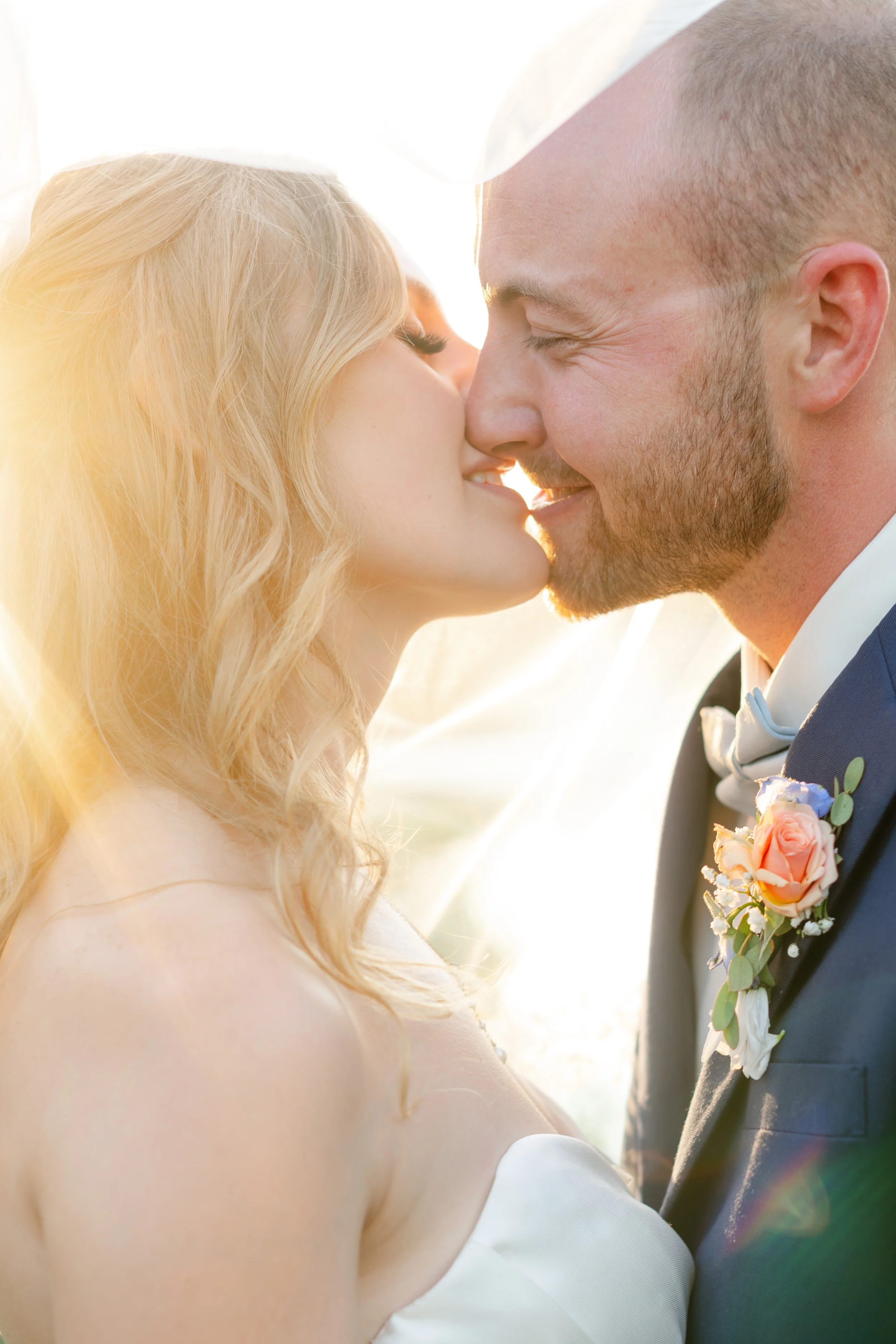 Bride and groom share a romantic kiss on their wedding day, sunlight shining softly around them.