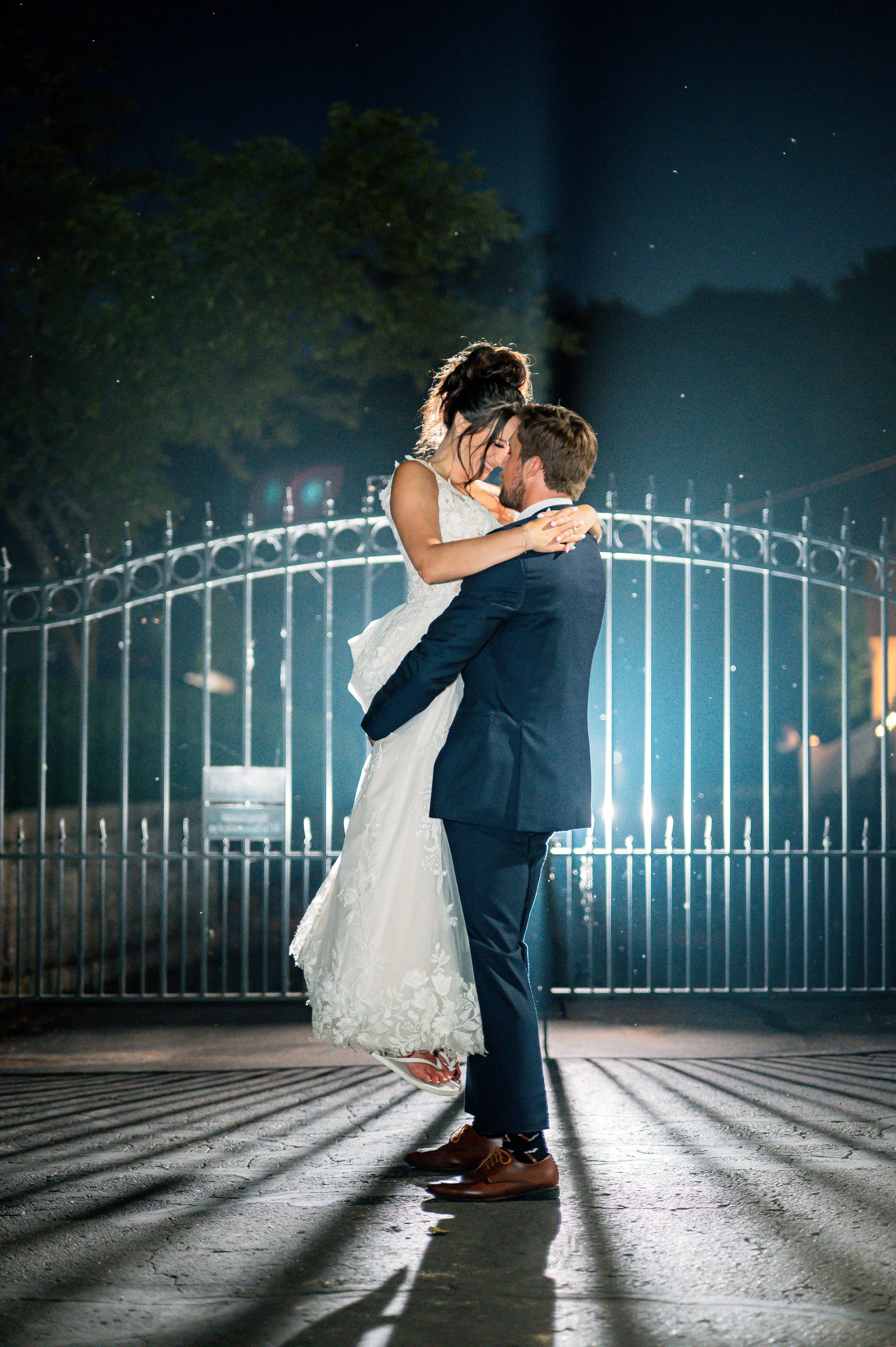 A bride and groom dancing at night outside, with a lit metal fence in the background and a tree overhead.