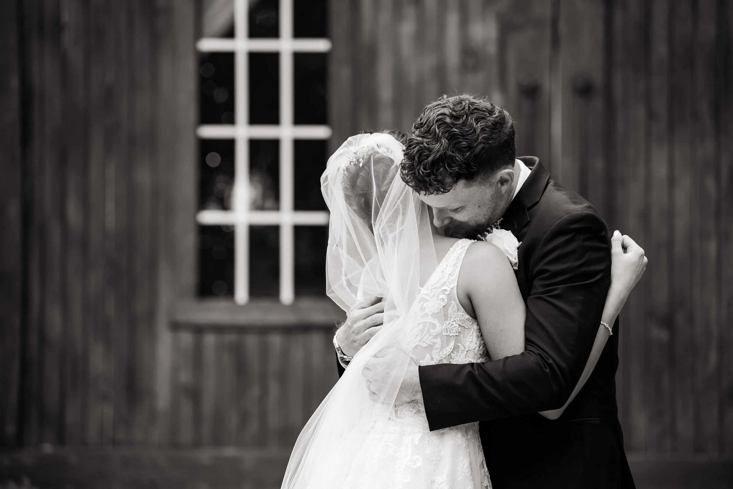 Black and white photograph of a bride and groom embracing each other closely on their wedding day, standing in front of a wooden building with a window.