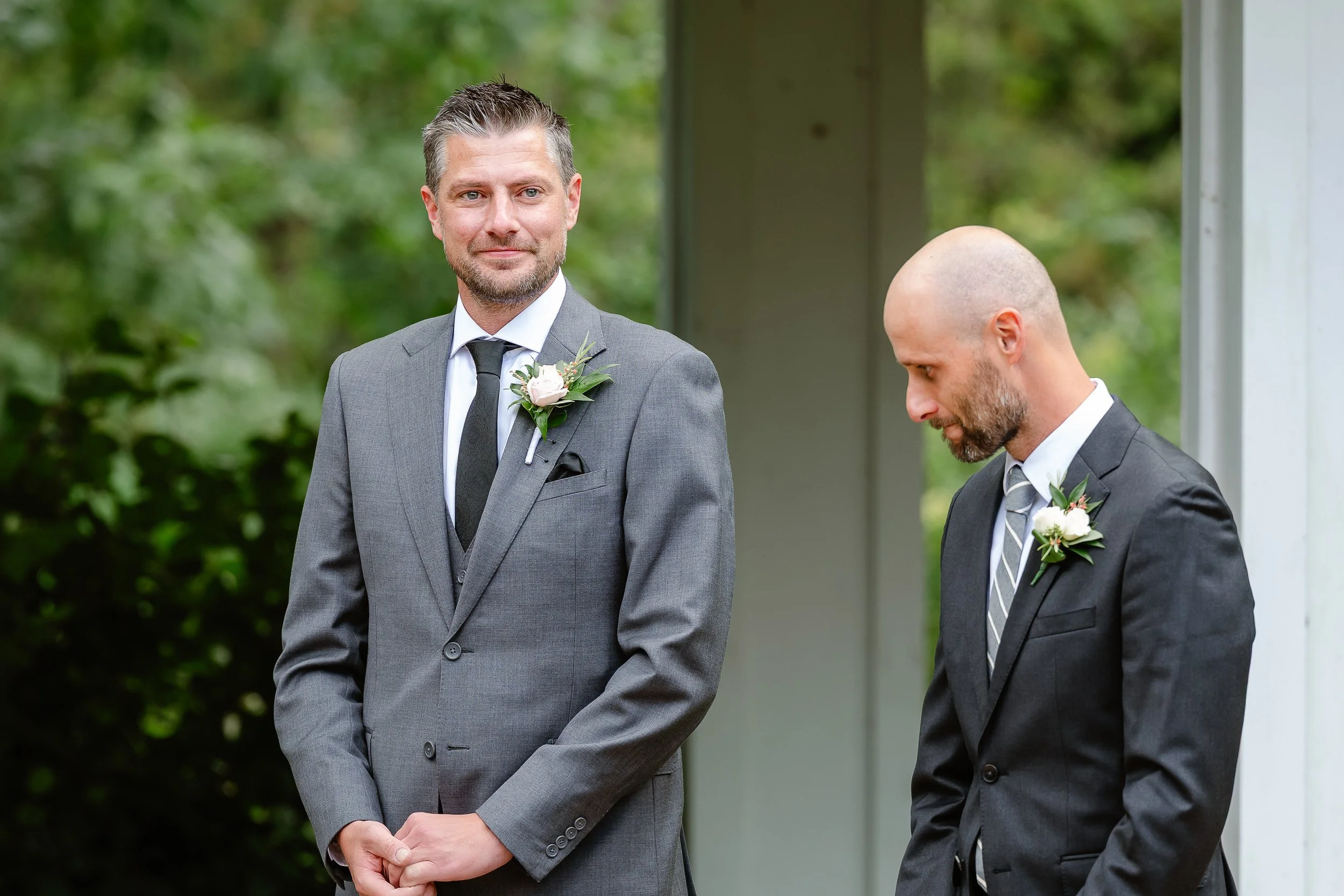Two men in suits with boutonnieres, standing outdoors, one looking at the camera and the other looking down.