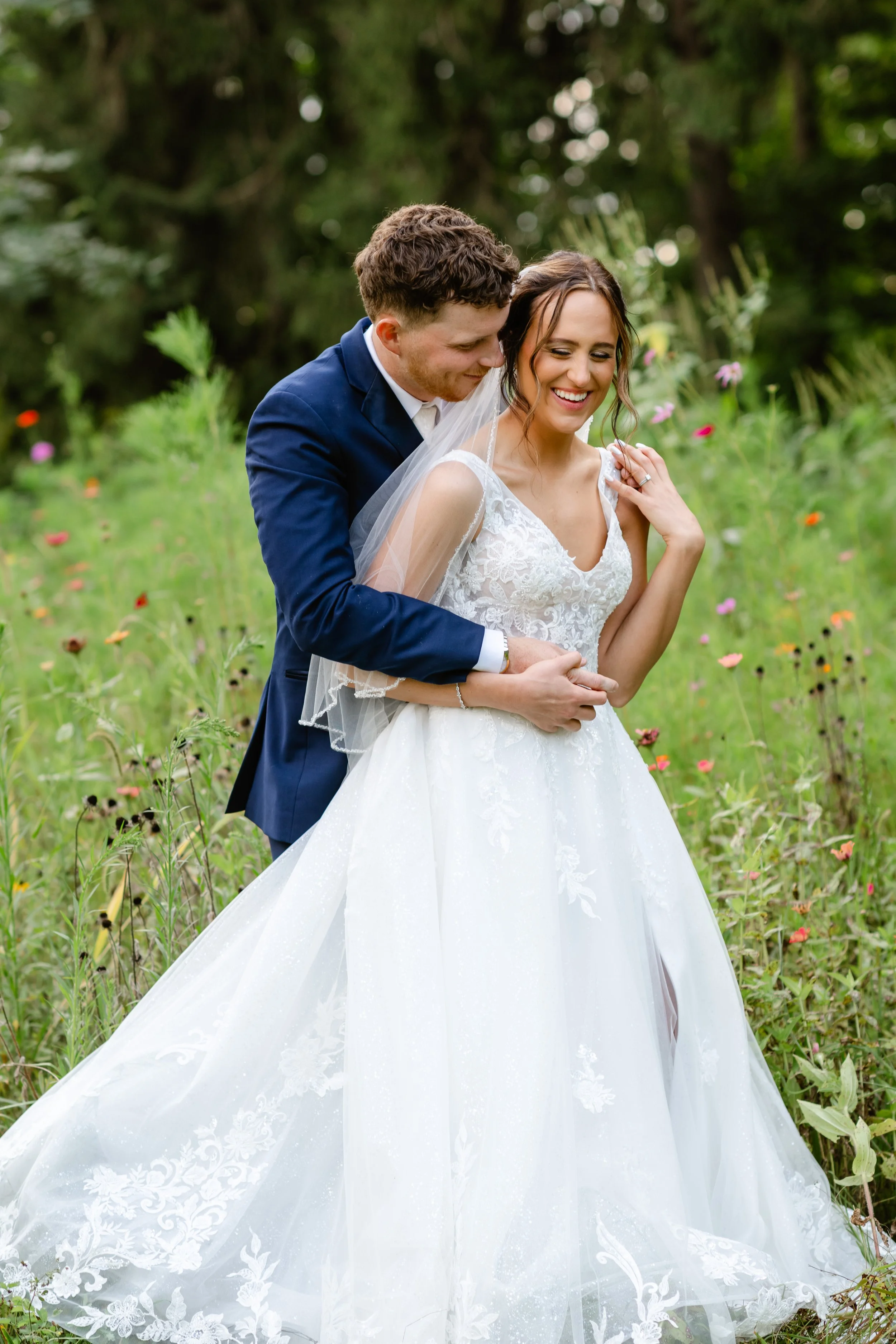 A smiling bride in a white wedding dress and a groom in a navy suit embracing in a field of flowers and greenery.