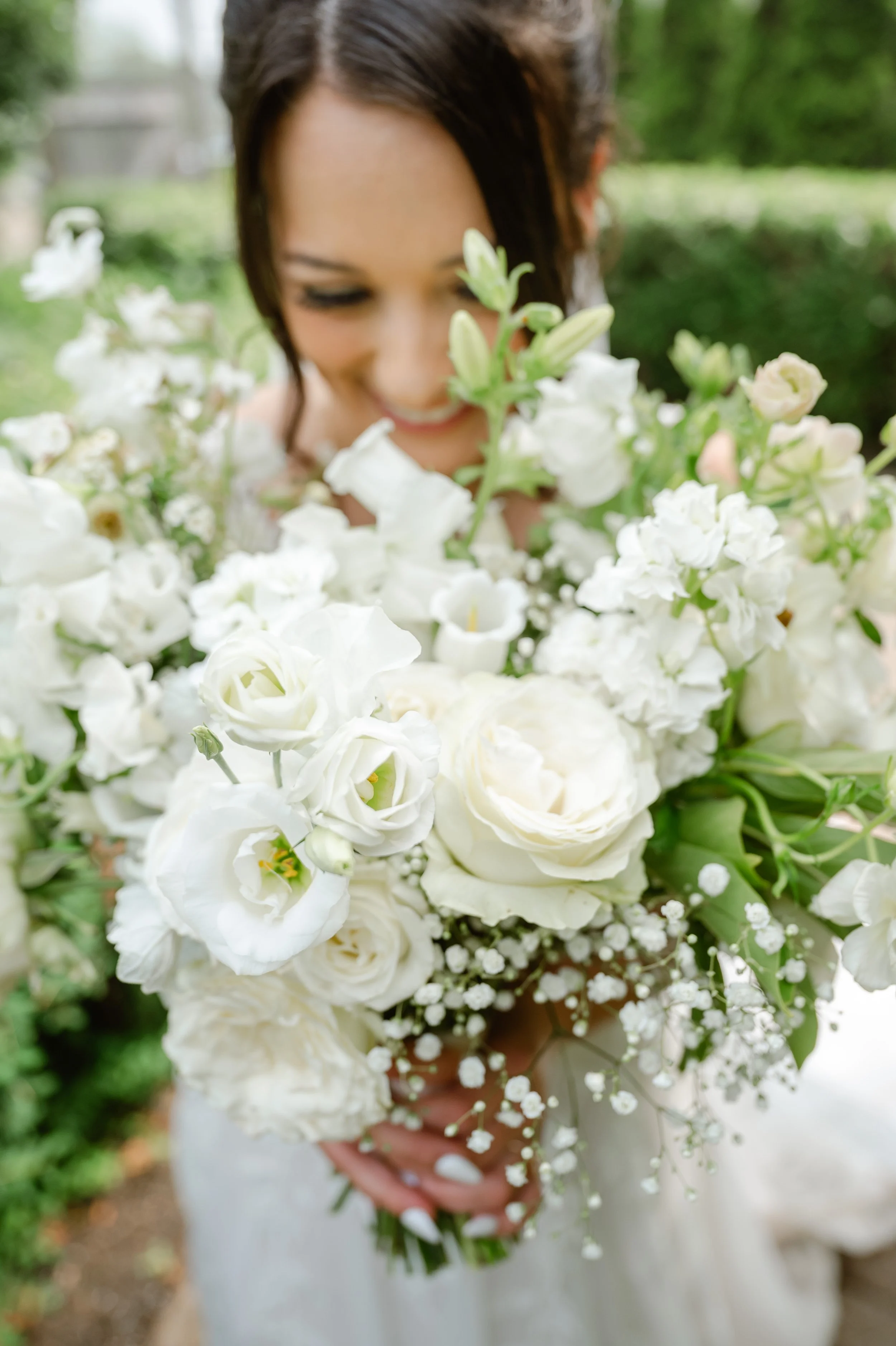 A woman in a wedding dress holding a large bouquet of white flowers, including roses and lisianthus, outdoors with green foliage in the background.