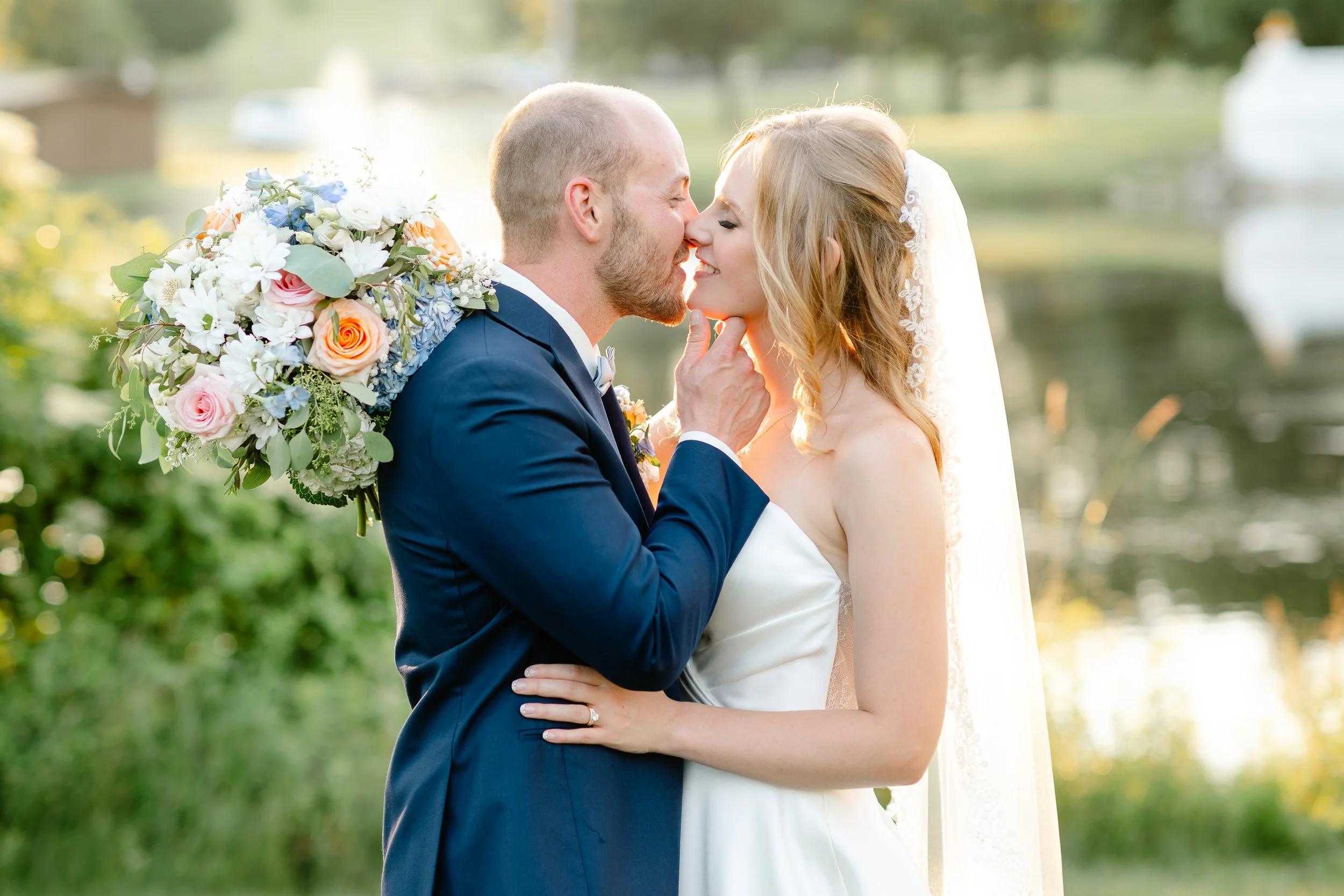 A newlywed couple sharing a kiss outdoors, the groom in a navy suit holding a large bouquet of flowers on his shoulder, while the bride in a white wedding dress and veil touches the groom's face, with a pond and trees in the background during sunset.