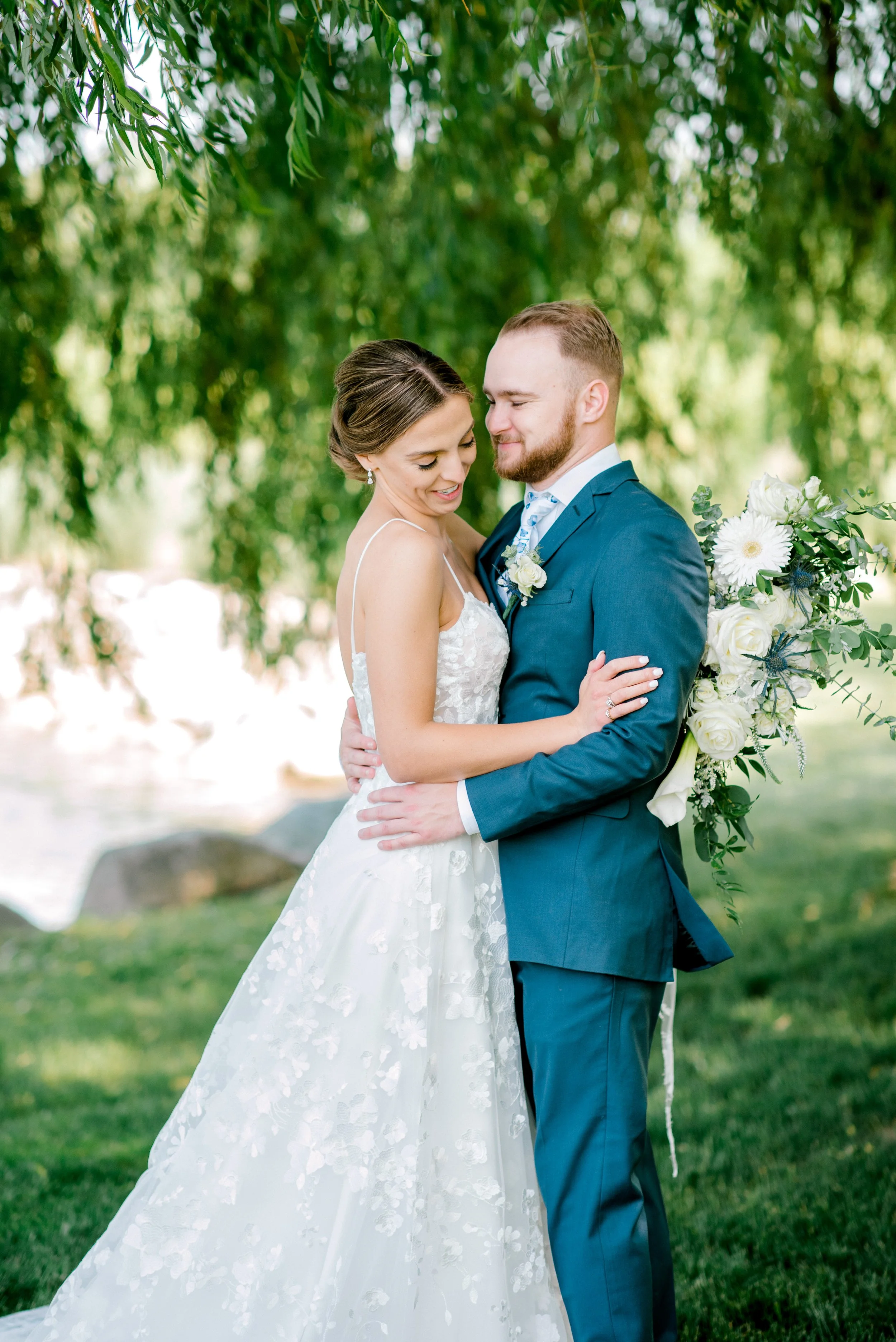 A bride and groom hugging outdoors, with trees and water in the background. The bride is in a white lace wedding gown, and the groom is in a blue suit holding a bouquet of white flowers.