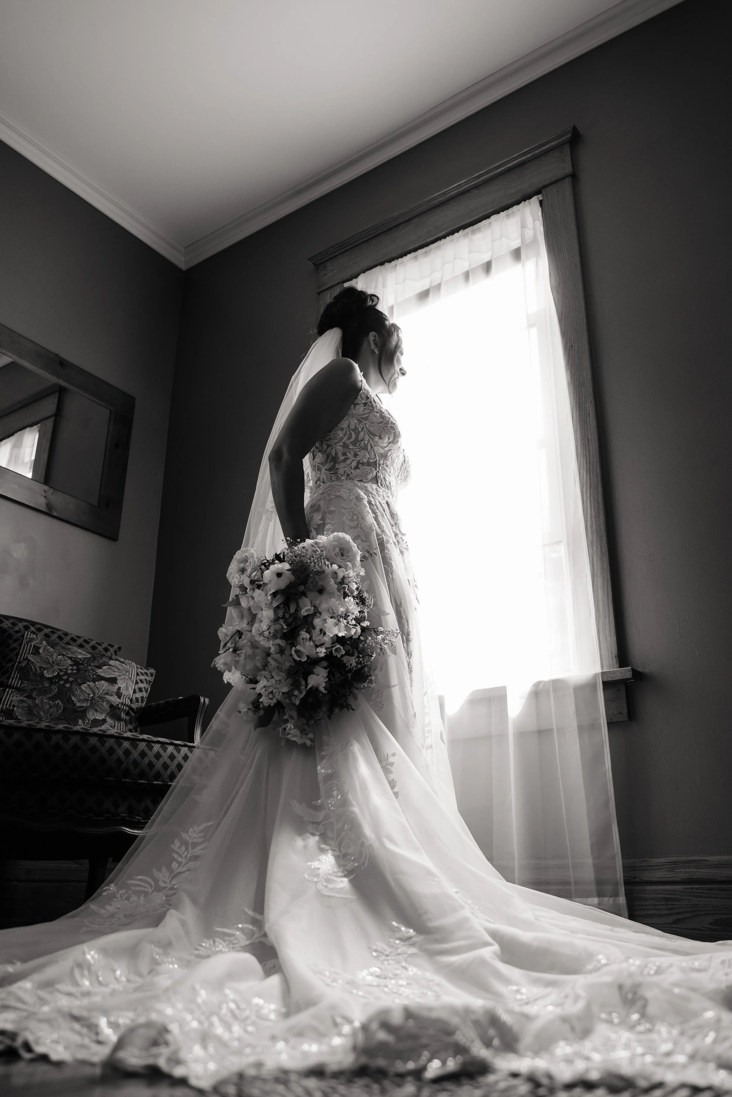 A bride in a wedding gown holding a bouquet, standing indoors near a window with curtains, in black and white.