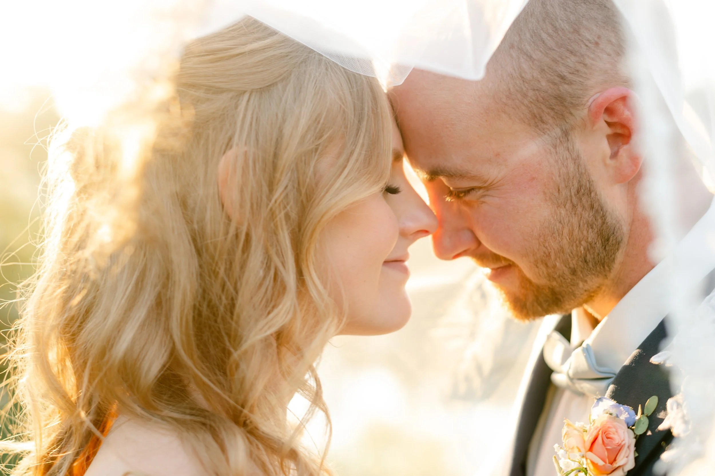 A bride and groom with their foreheads touching, smiling gently, outdoors during sunset, with sunlight shining through a veil.