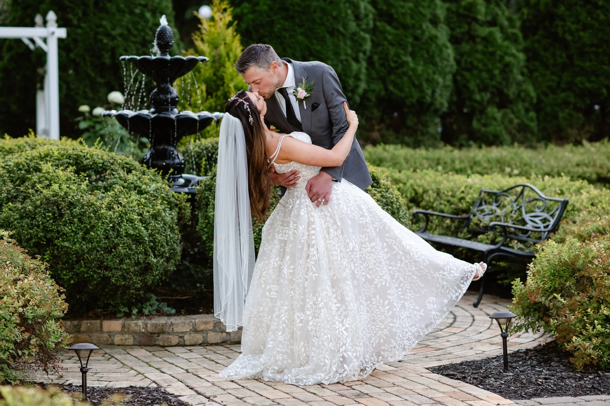 A newlywed couple sharing a kiss outdoors near a fountain, with greenery and garden benches in the background.