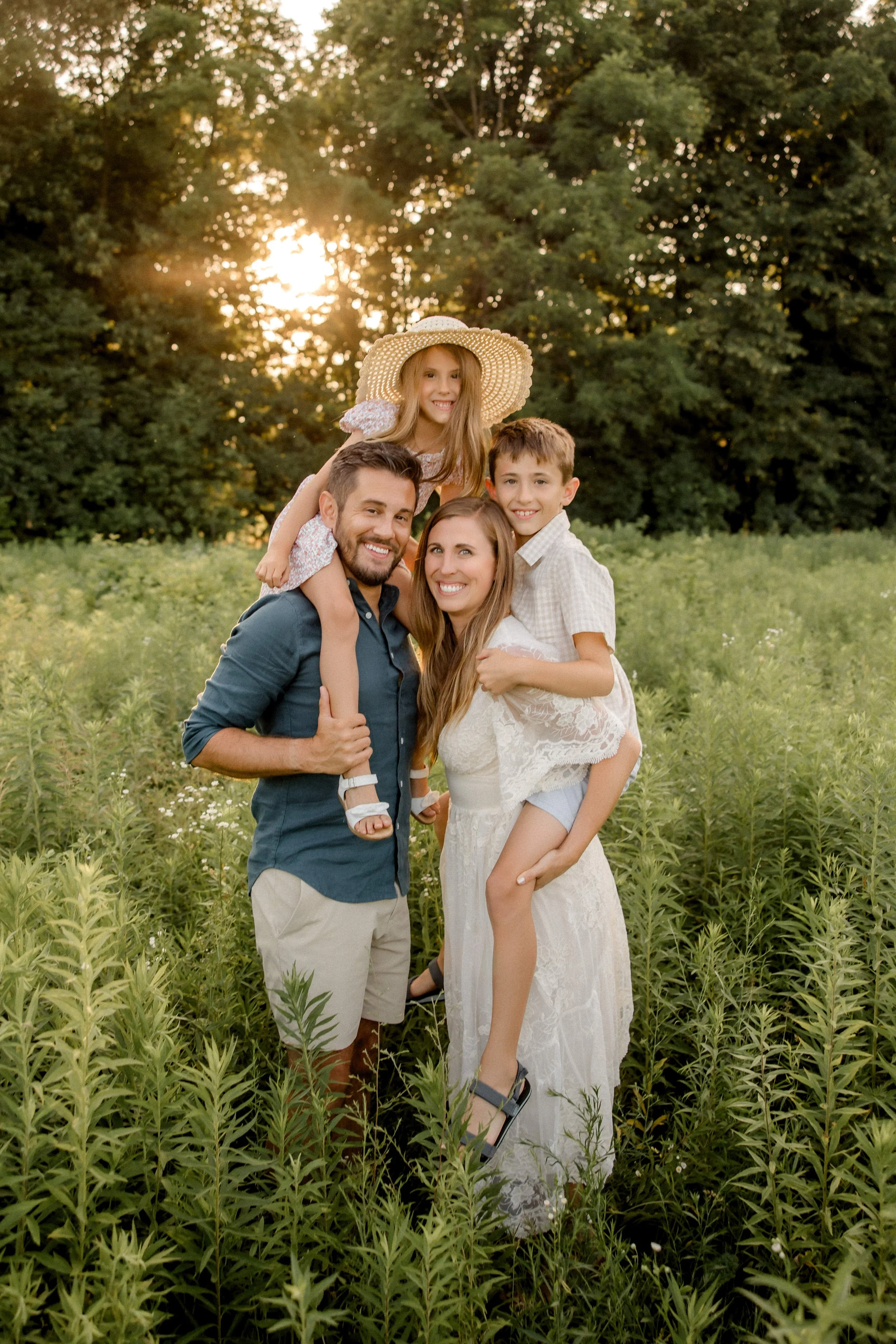 A family of four in a field with green plants and trees, smiling at the camera during sunset. The father is carrying his daughter on his shoulders, the mother is holding their son, and they are dressed in casual summer clothes.