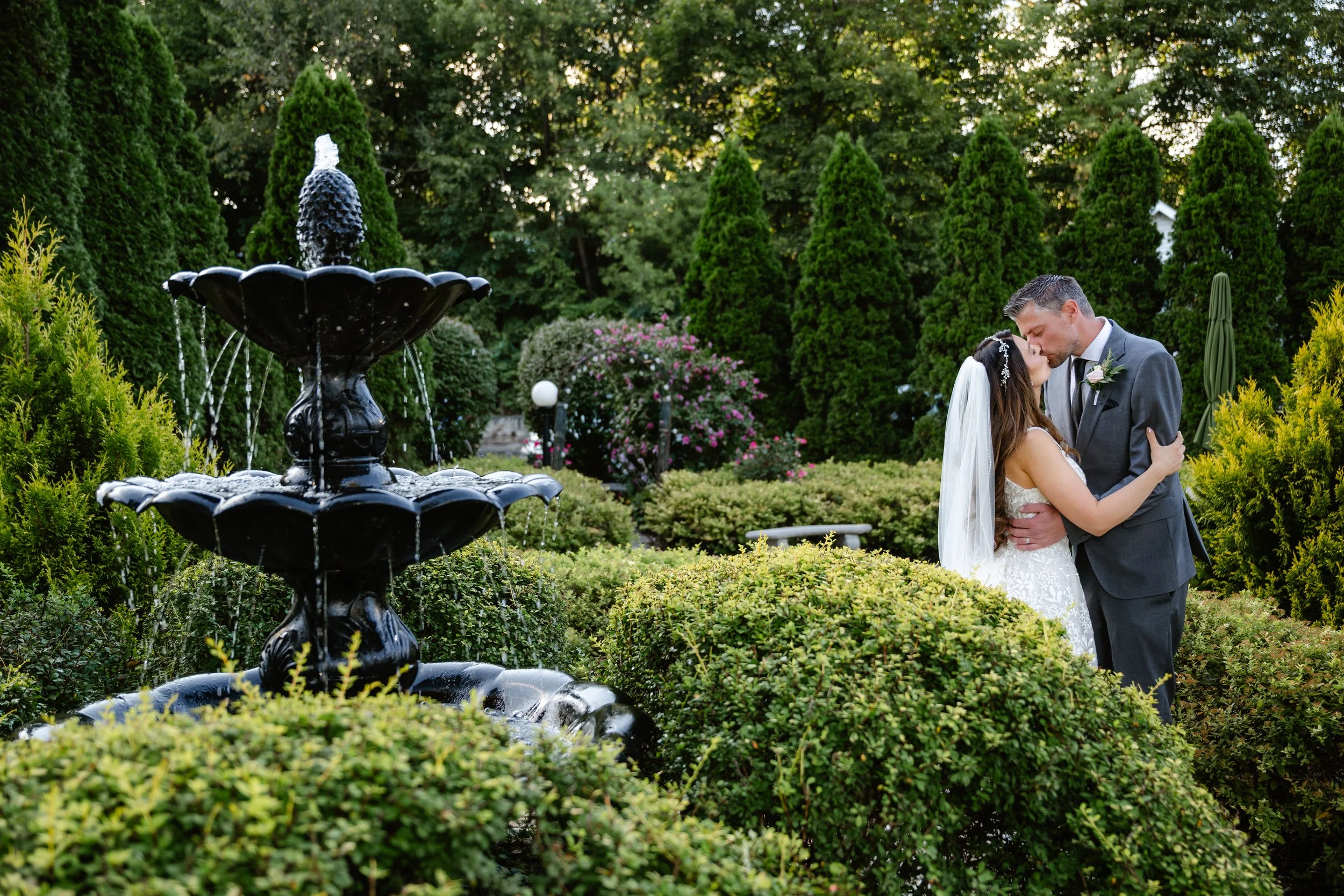 A bride and groom sharing a kiss in a lush garden with a black fountain in the foreground and green trees in the background.