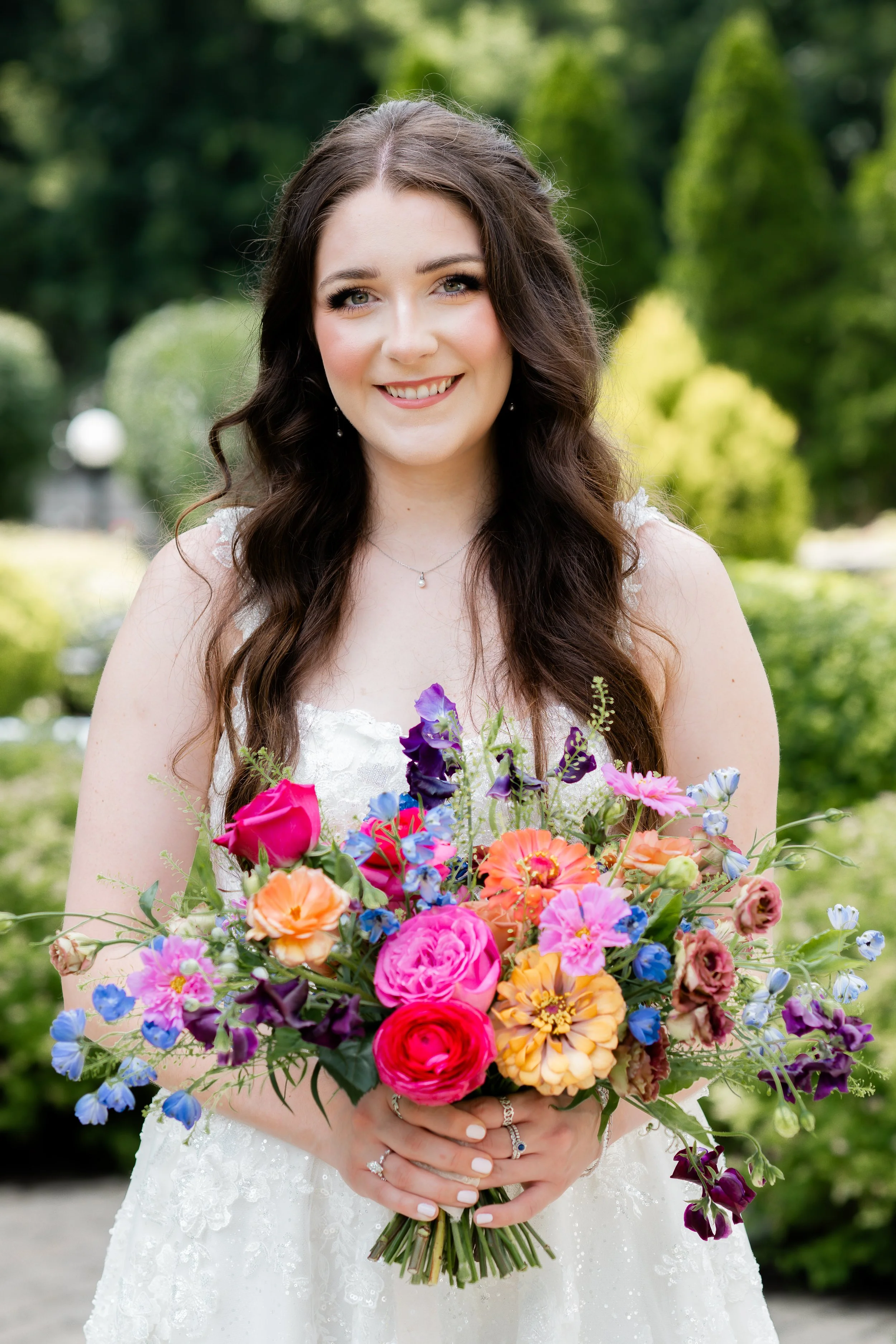 A smiling woman in a wedding dress holding a colorful bouquet of flowers outdoors in a garden.