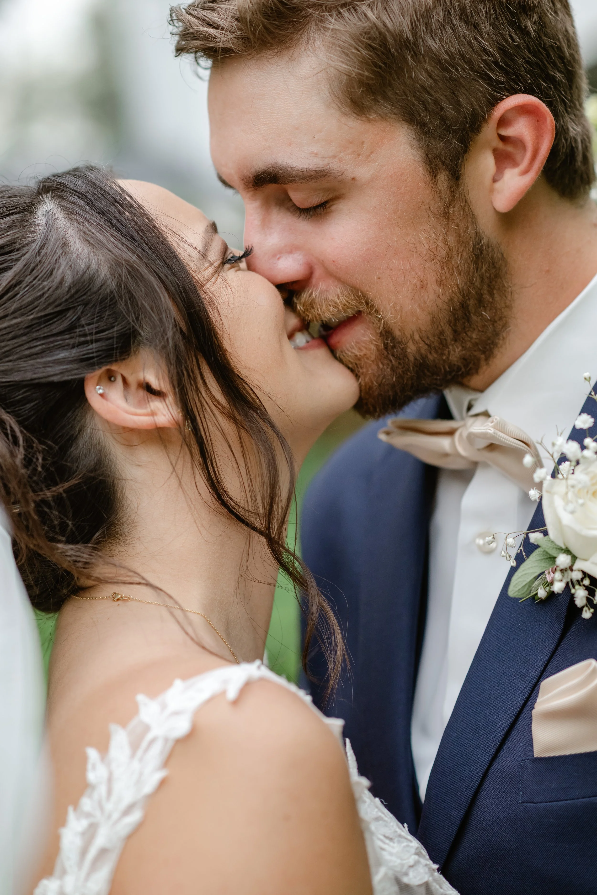 A newlywed couple sharing a kiss, with the bride wearing a white lace dress and the groom in a navy suit with a beige bow tie and boutonniere.