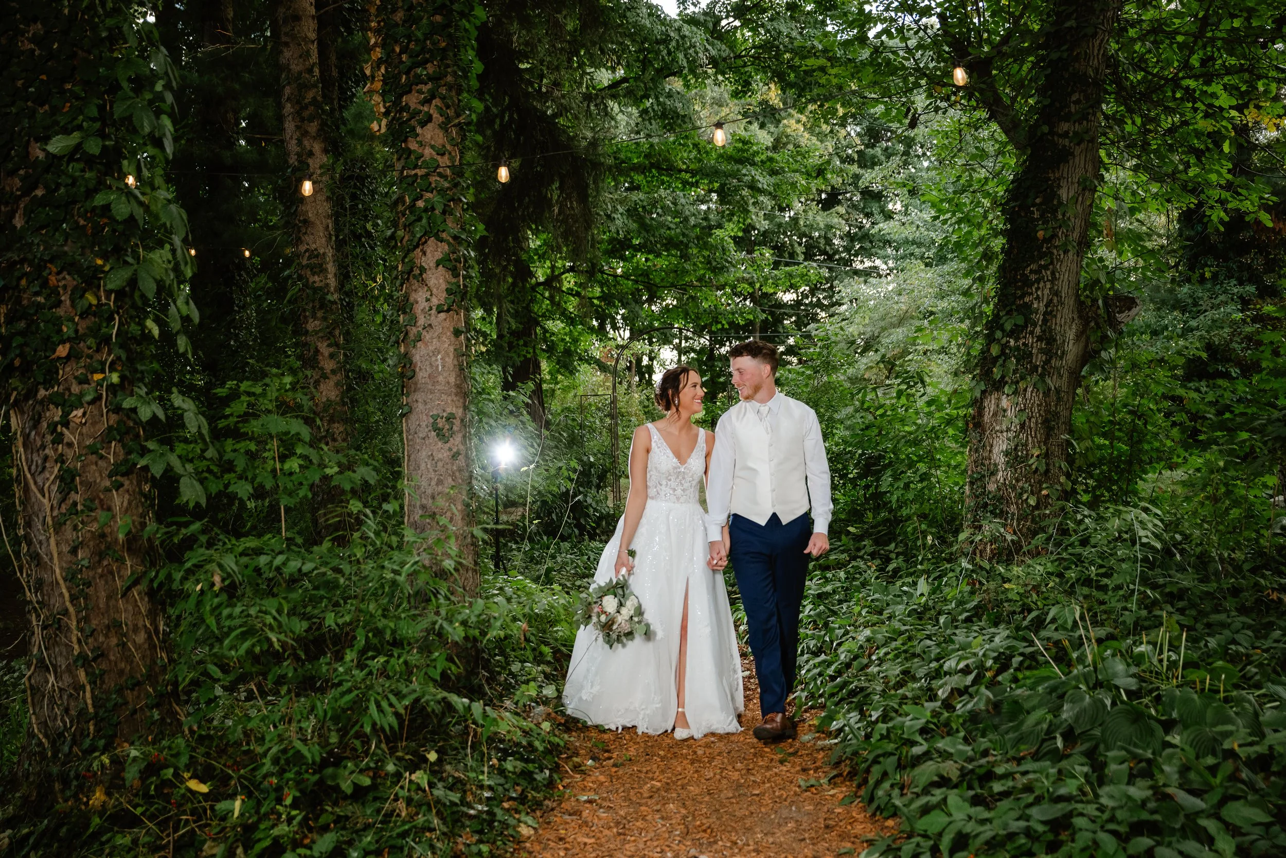 Wedding couple holding hands and walking in a lush, green forest with string lights hanging overhead.