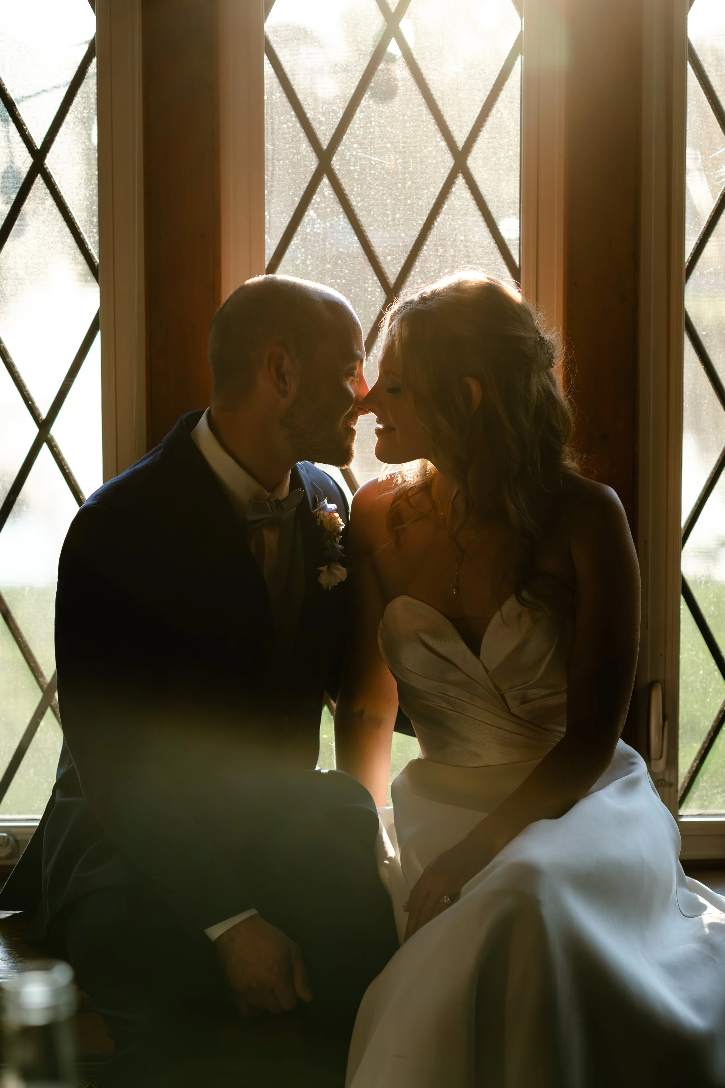 A bride and groom sit close together by a window, touching foreheads, with backlit sunlight creating a silhouette effect.