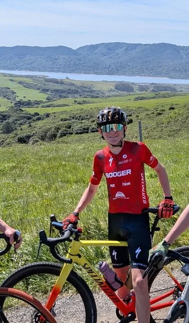 A woman in a red cycling jersey and black shorts stands with her yellow and black road bike in a grassy field. She wears a black helmet, reflective sunglasses, and red gloves, smiling at the camera. The background features rolling green hills, a body of water, and mountains under a partly cloudy sky.