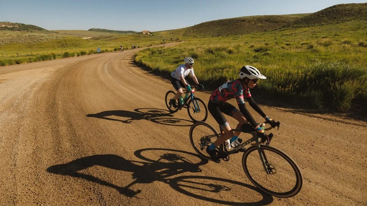 Two cyclists riding mountain bikes on a gravel road in a scenic, hilly landscape with green grass and distant hills.