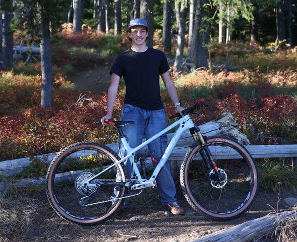 Person standing outdoors in a wooded area, holding a mountain bike.