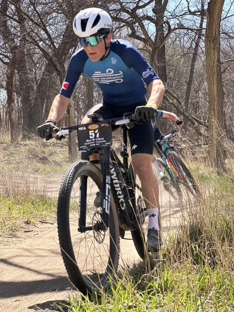 Cyclist riding a mountain bike on a dirt trail through a wooded area, wearing a blue jersey, white helmet, and sunglasses.
