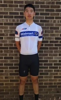 A young man in a Walmart cycling jersey and black shorts standing against a brick wall.