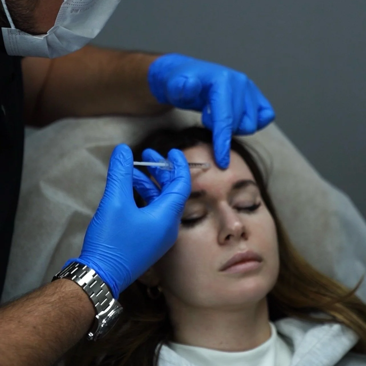 A woman receiving a cosmetic injection in her forehead while lying down, with a healthcare professional wearing blue gloves administering the shot.