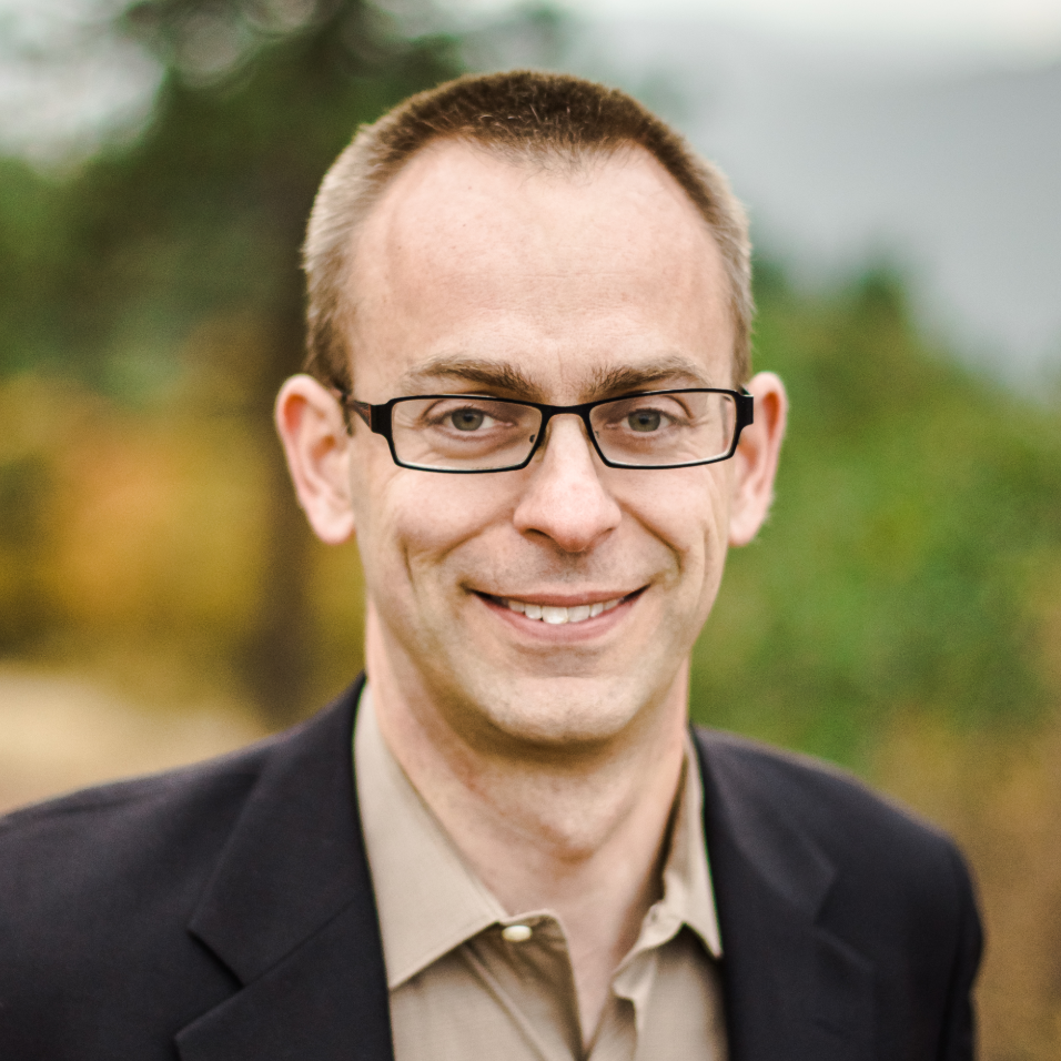 Travis is a smiling man with short brown hair and dark rectangular glasses.