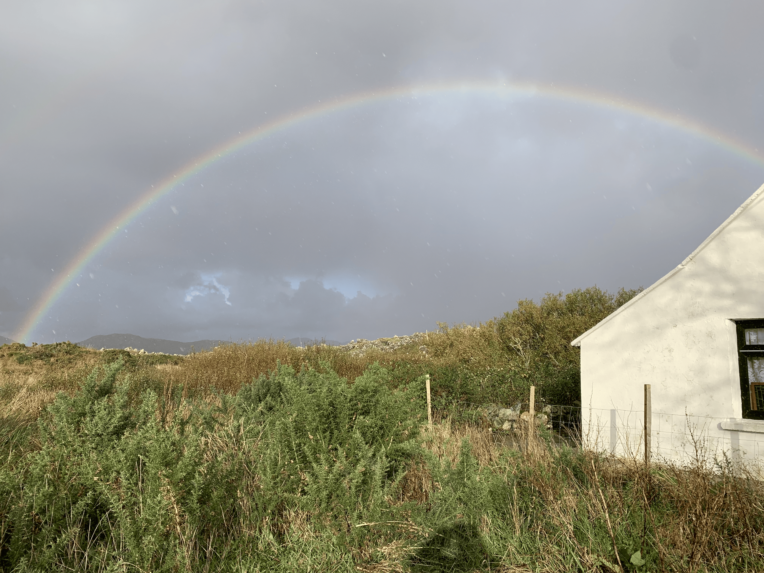 A rainbow arching across a cloudy sky over a rural landscape with a small white house on the right and trees and hills in the background.