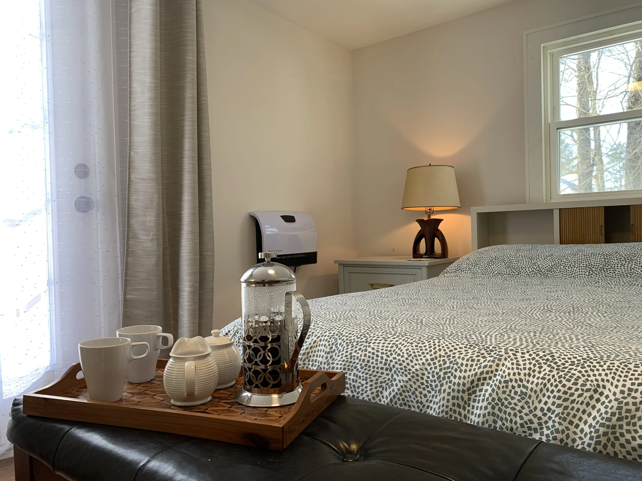 A cozy bedroom with a bed featuring a patterned duvet, a wooden bedside table with a lamp, and a window with curtains. A tray on the bed holds a collection of white coffee mugs and decorative pitchers.