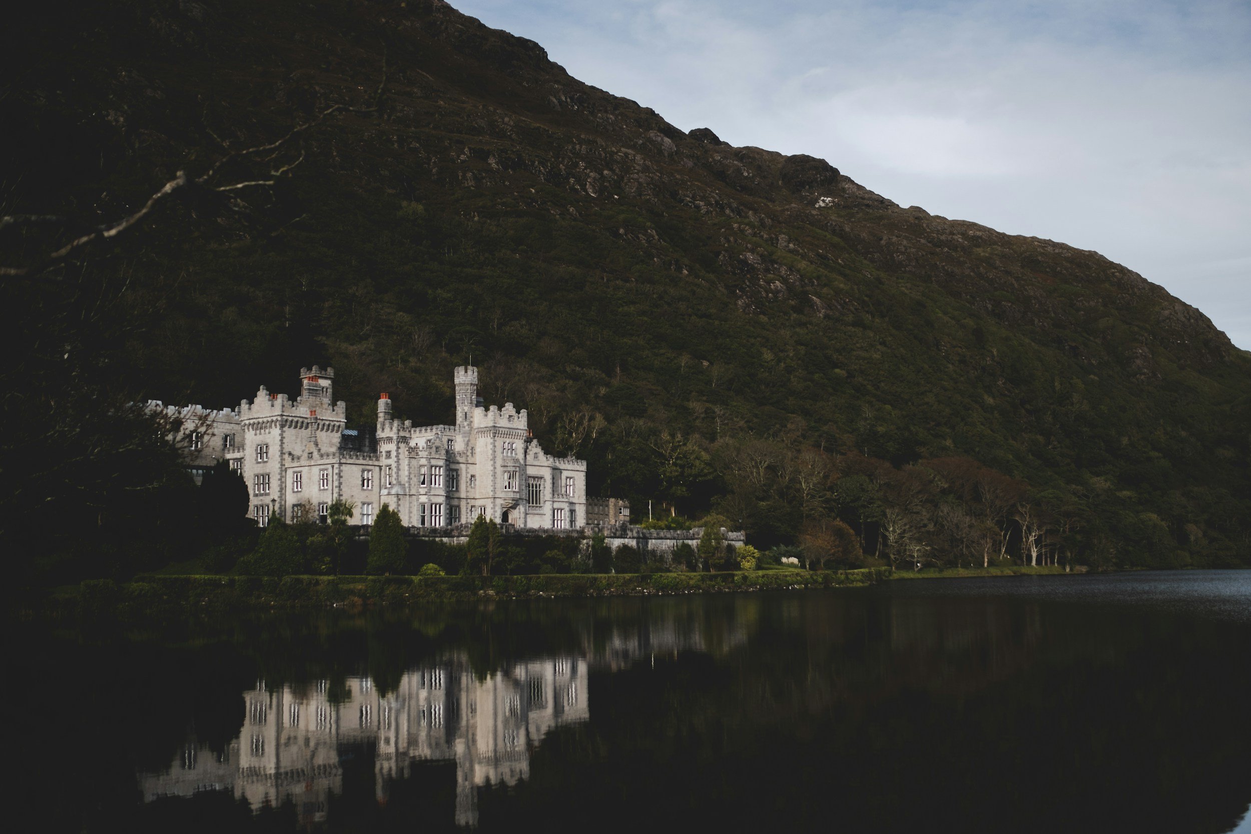 A large, white castle-style mansion with turrets and towers, situated at the foot of a dark, wooded mountain, with its reflection visible in a calm body of water in the foreground.