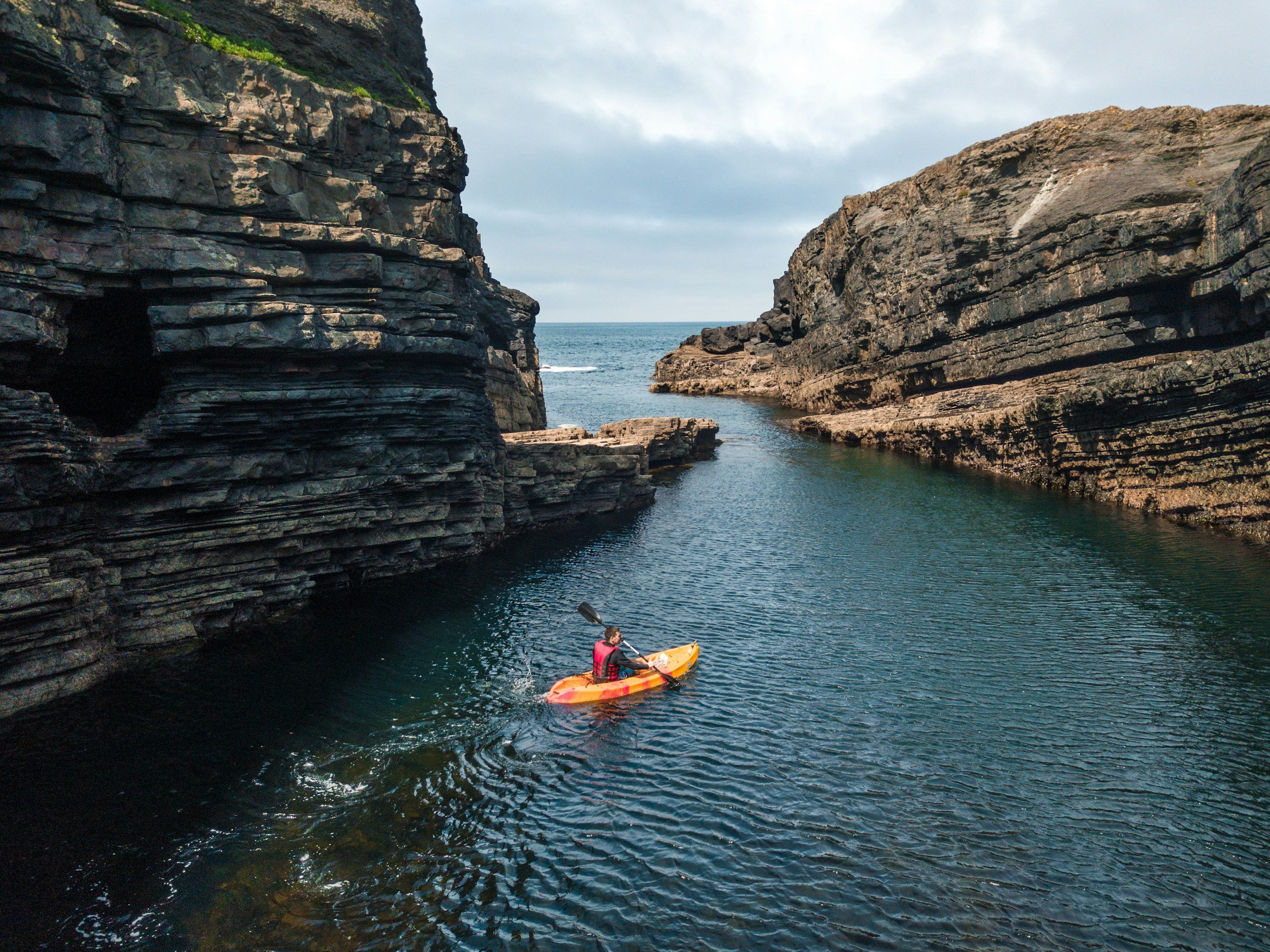 A person kayaking in a narrow water passage between large rocky cliffs under a cloudy sky.