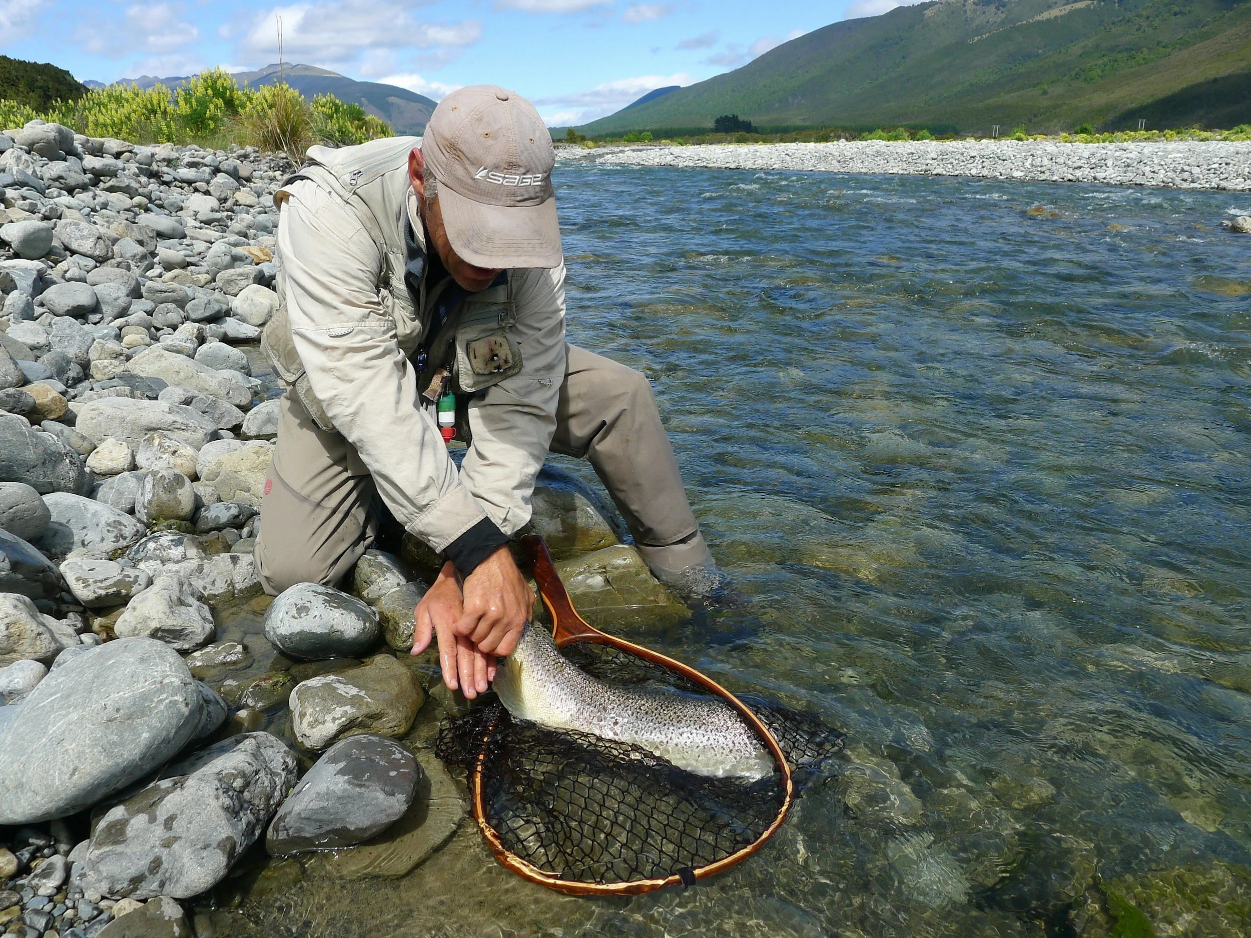 A person in outdoor clothing and a hat is kneeling on a rocky riverbank, holding a large fish over netted water.