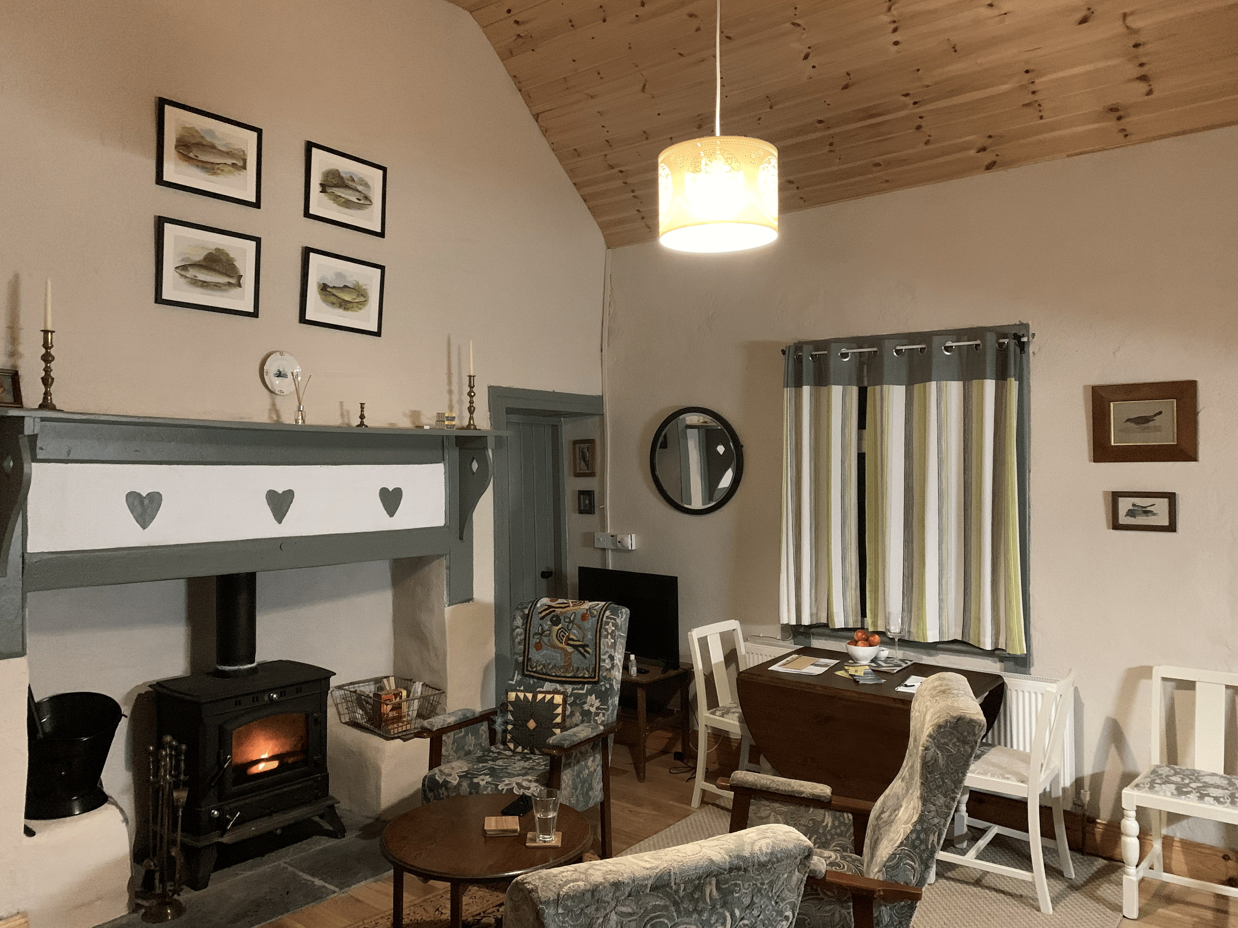 Living room with a wood stove, framed pictures on the wall, a curtain with stripes, various chairs and a table with a bowl of fruit.