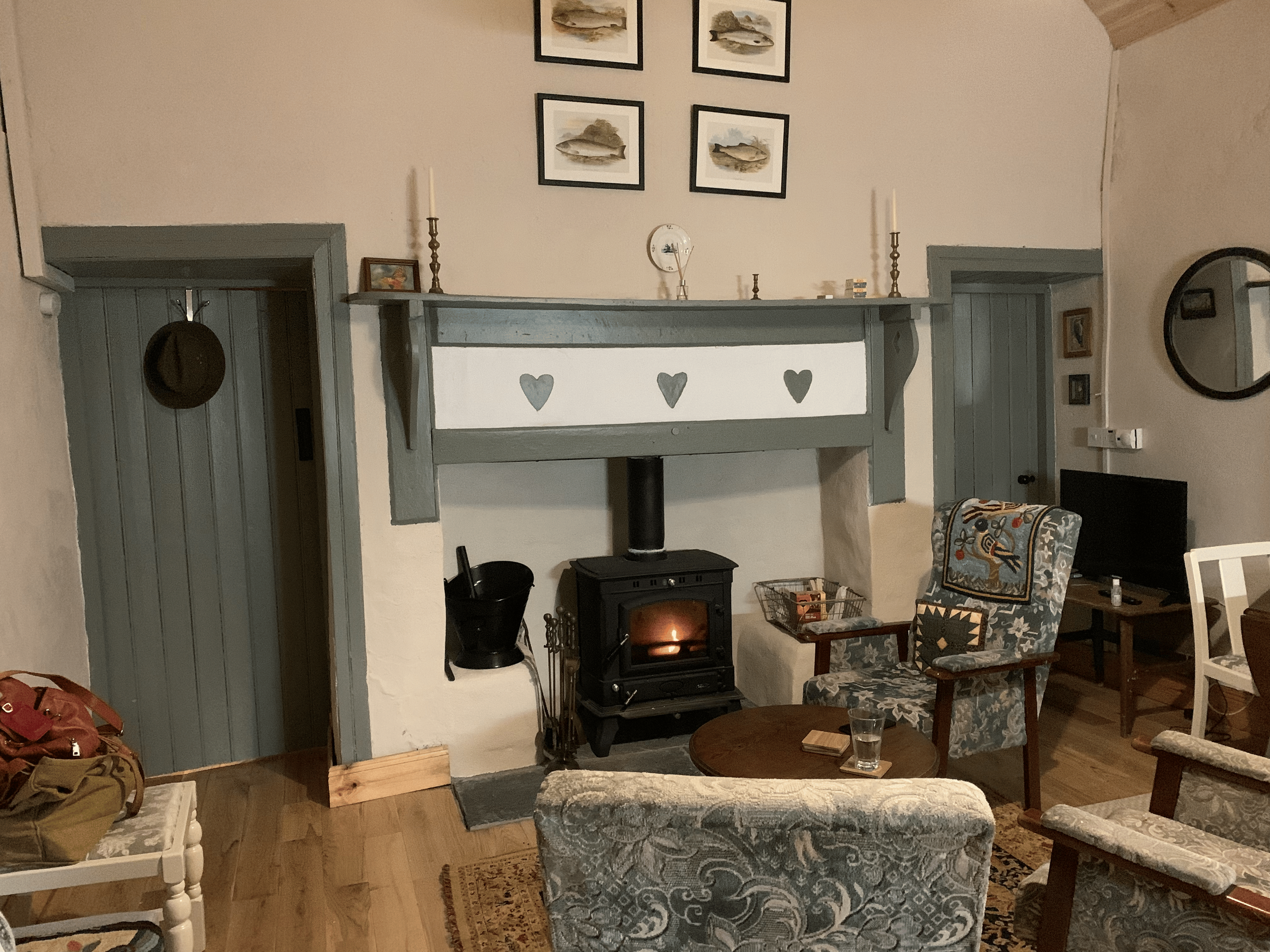 Living room with a wood stove, framed pictures of fish on the wall, a gray fireplace mantel with candles and small decorative items, patterned armchairs, a clock on the wall, and various furniture and items.