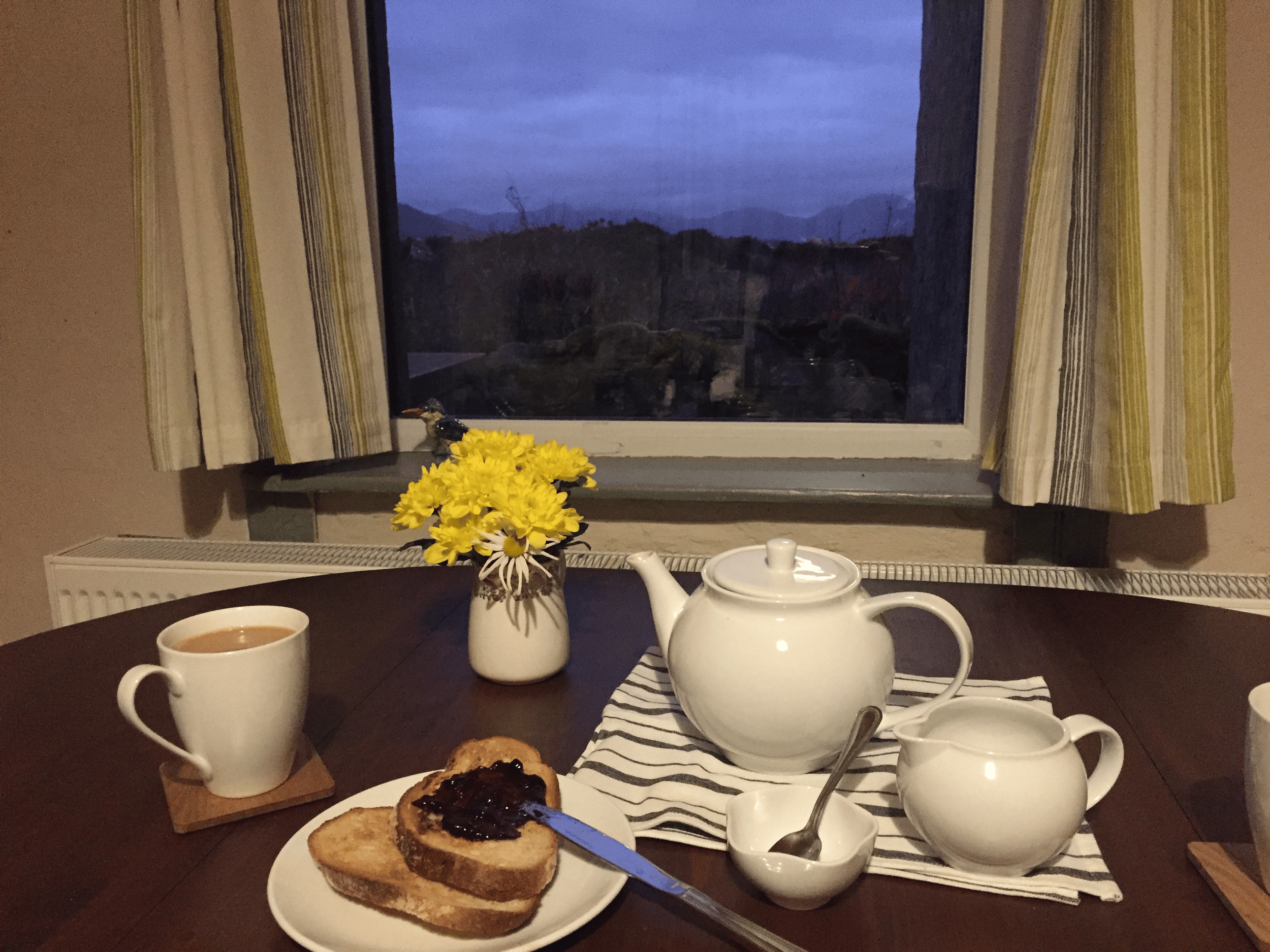 A cozy breakfast scene viewed through a window showing mountains and cloudy sky at dusk. Inside, a dark wooden table has a white teapot, a white mug with tea or coffee, a small bowl with cream, and a plate with toasted bread and jam. There is a vase 