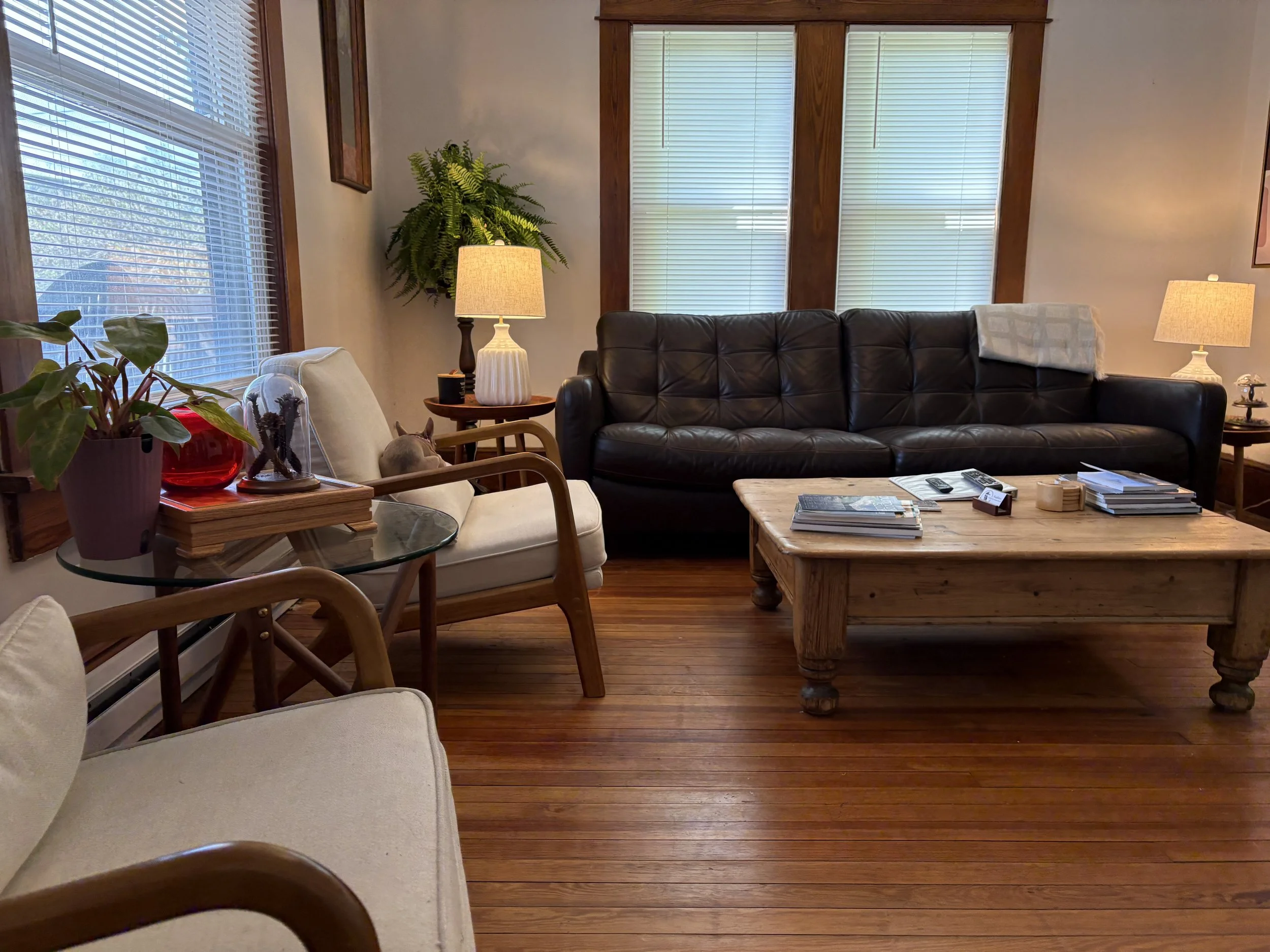 Living room with wooden floors, a black leather couch, beige armchair, side table with a lamp, and large windows with blinds. A small dog is lying on the armchair, and various decorative items and magazines are on the coffee table.