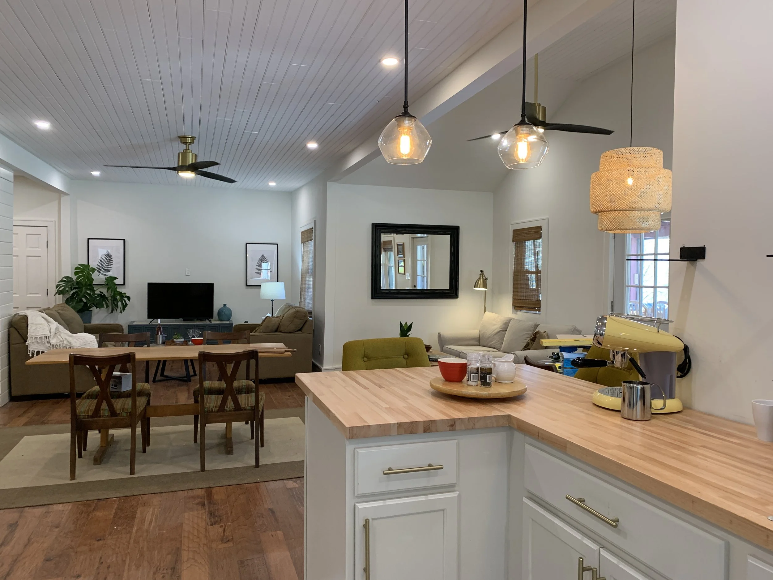 Open-concept living and kitchen area with hanging pendant lights, ceiling fans, a dining table, and various furniture pieces, decorated with framed artwork and houseplants.